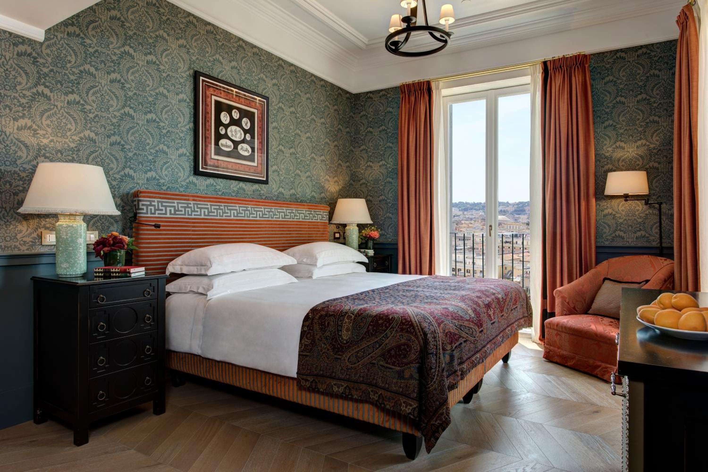 Ornate paisley wallpaper and rich burgundy textiles create an intimate sanctuary, while French doors frame Rome's terracotta rooftops beyond the private balcony.