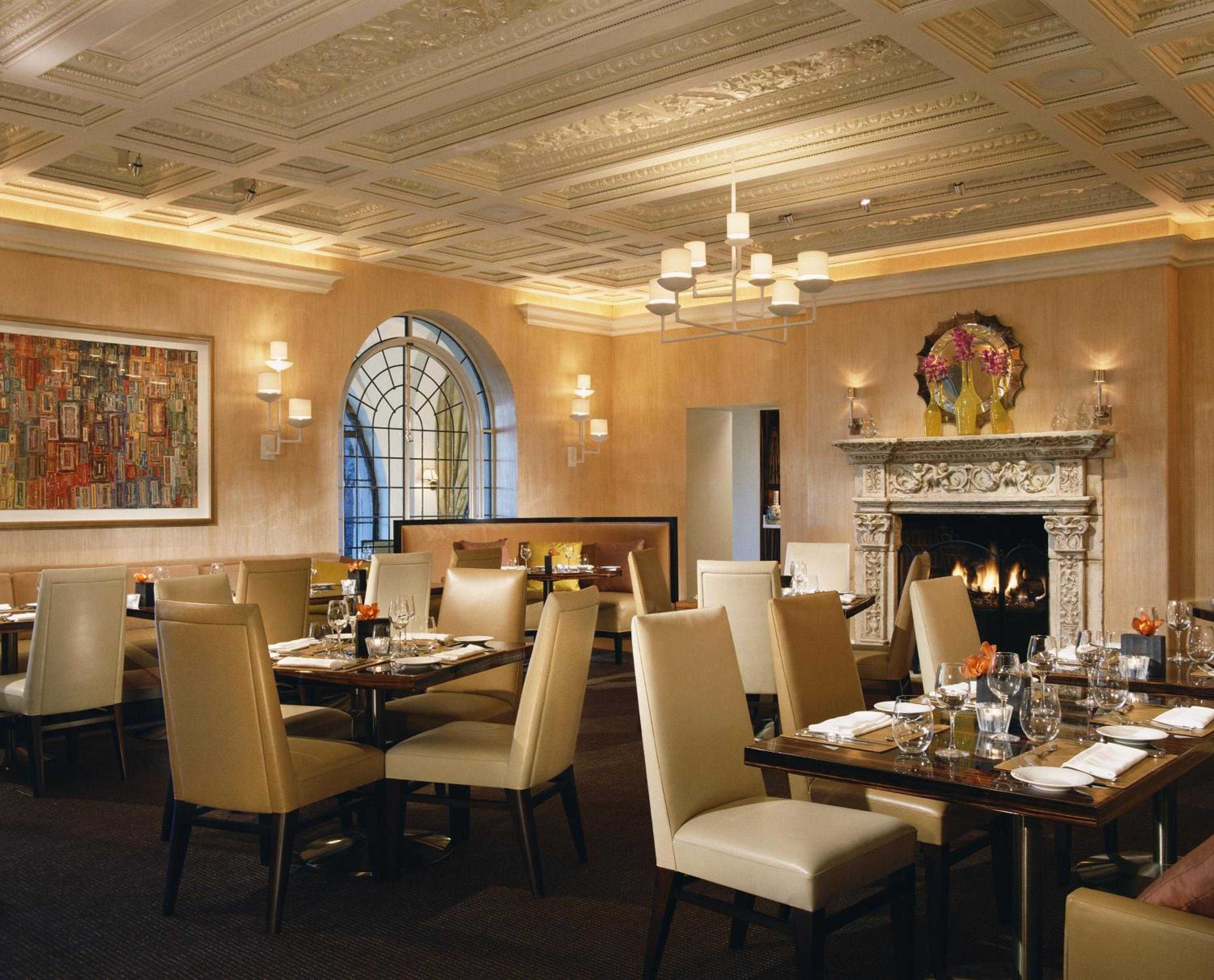 Coffered ceilings and warm amber lighting create an intimate atmosphere in the hotel's signature restaurant, where cream leather chairs gather around polished wood tables beside a crackling fireplace.
