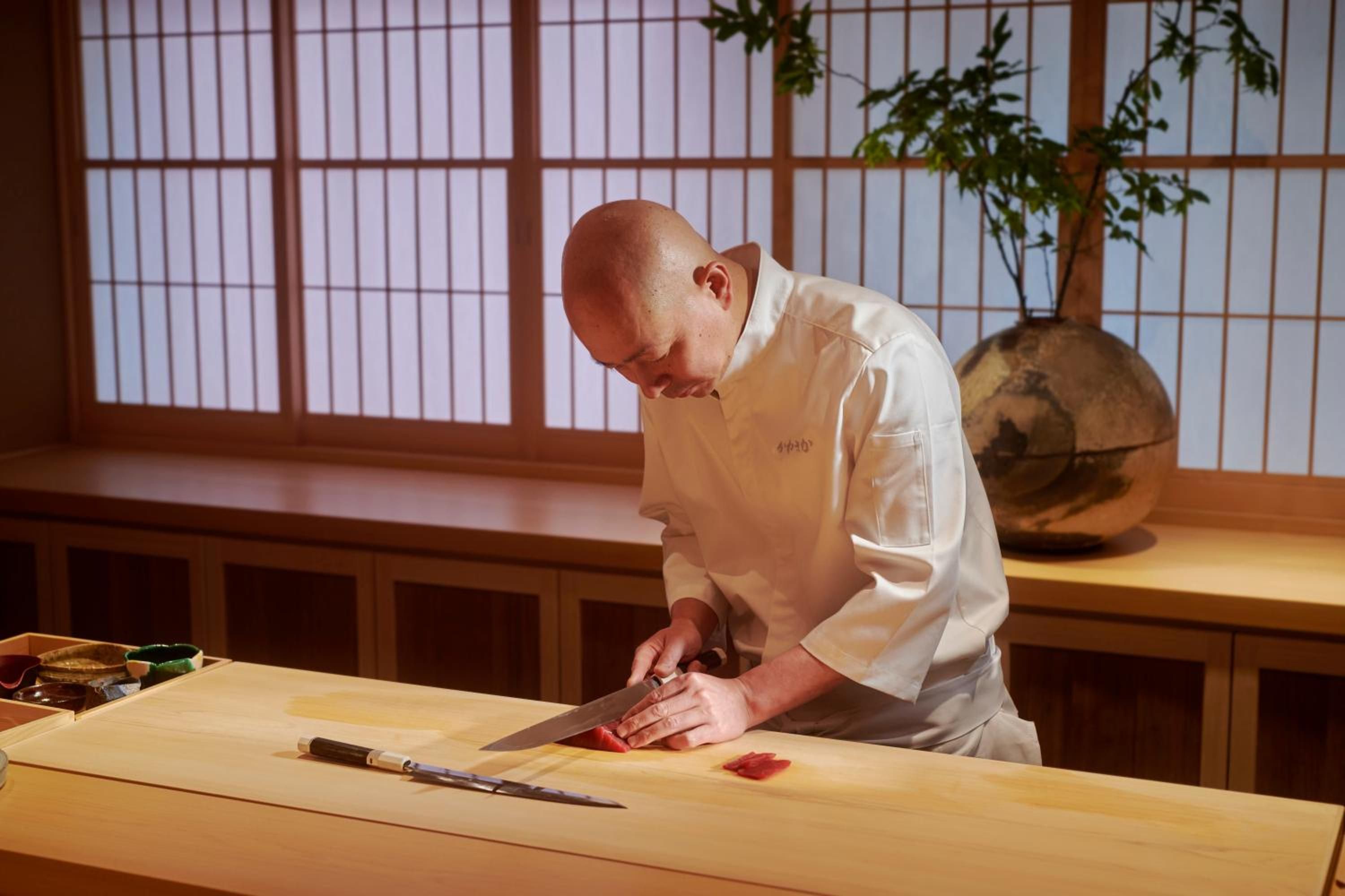 Master sushi chef's blade glides through pristine fish at the counter, each precise cut a meditation in Japanese culinary artistry behind traditional shoji screens.