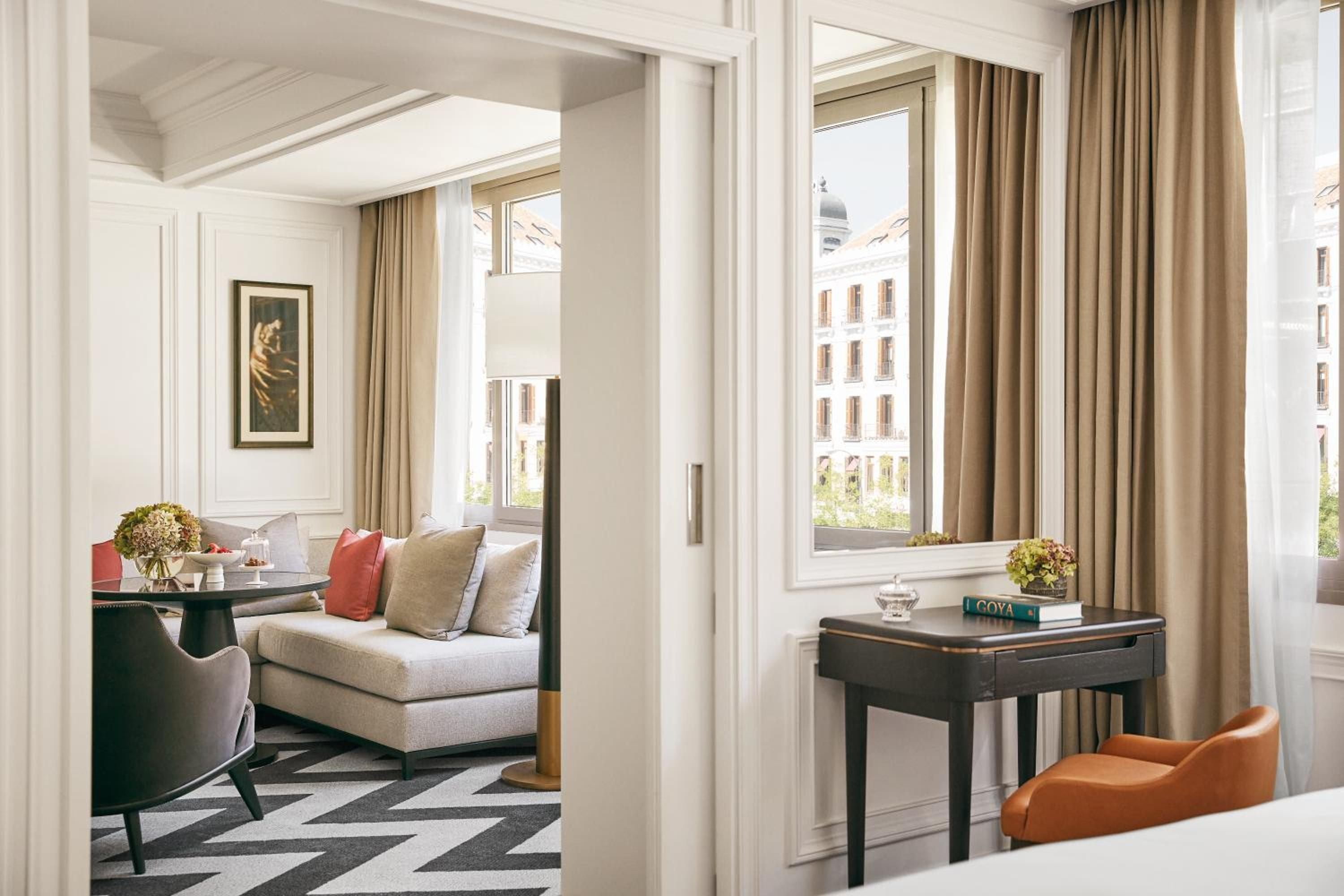 Elegant proportions define this corner suite, where coral accents warm cream furnishings and French doors frame Madrid's historic rooftops beyond flowing silk curtains.