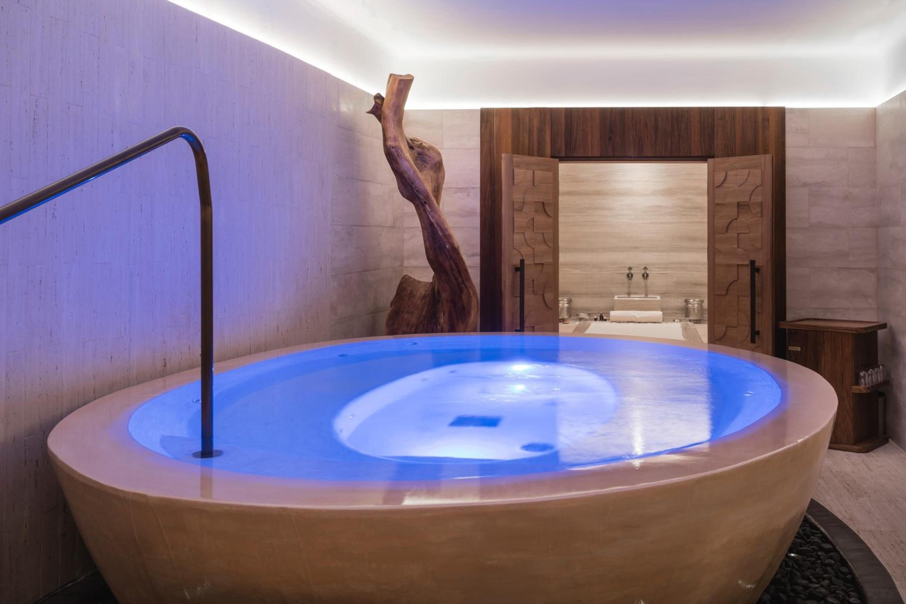 Illuminated by ethereal blue light, this sculptural soaking tub transforms the spa into a sanctuary where natural wood, stone textures, and artistic elements create pure tranquility.