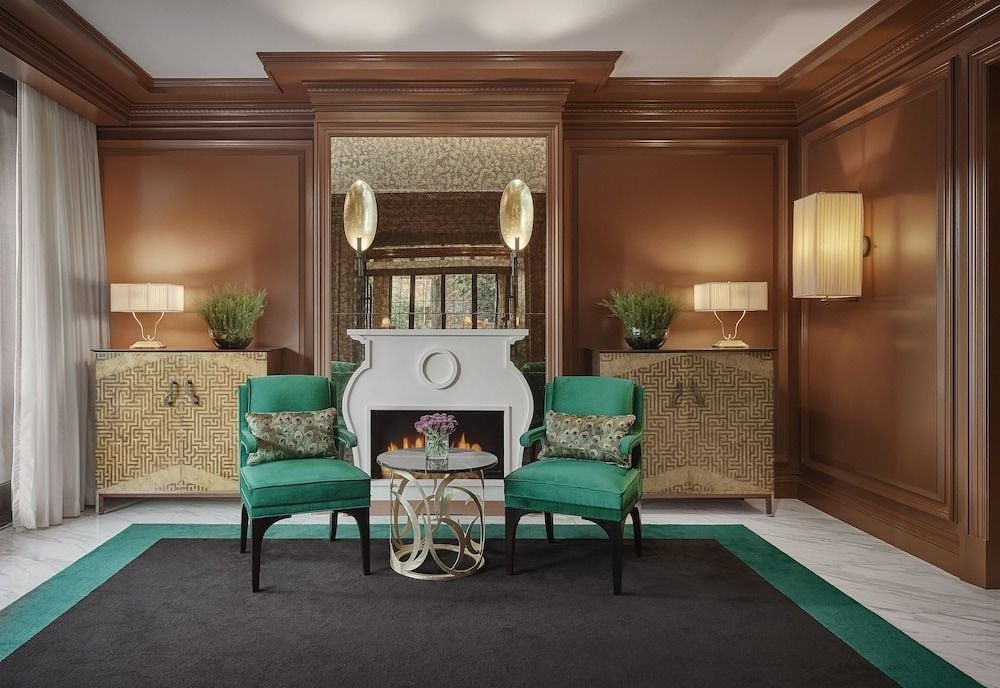 Rich walnut paneling frames emerald velvet chairs in this sophisticated seating area, where golden pendant lights cast warm shadows across geometric-patterned screens and marble fireplace details.