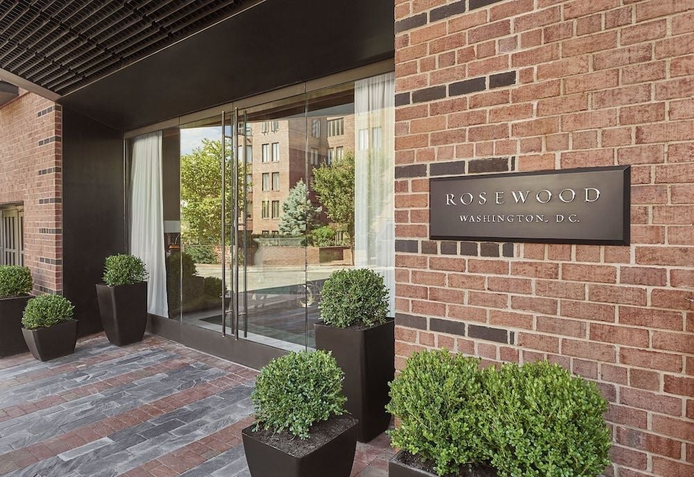 Warm brick facades and sleek glass doors create an inviting Georgetown entrance, where manicured boxwood topiaries in charcoal planters signal refined luxury ahead.