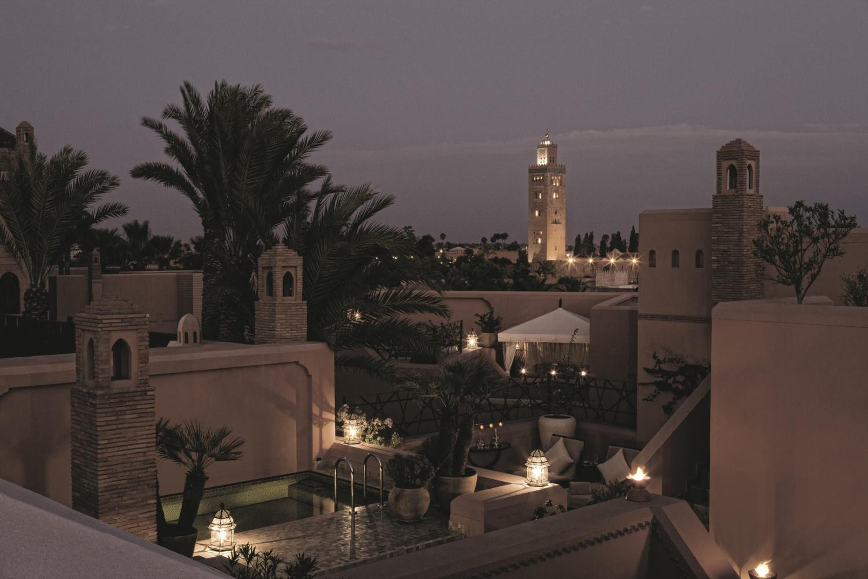 Illuminated terraces cascade beneath Marrakech's iconic minaret as twilight settles over Royal Mansour's rooftop riads, their lantern-lit courtyards glowing like jeweled boxes against the dusky sky.