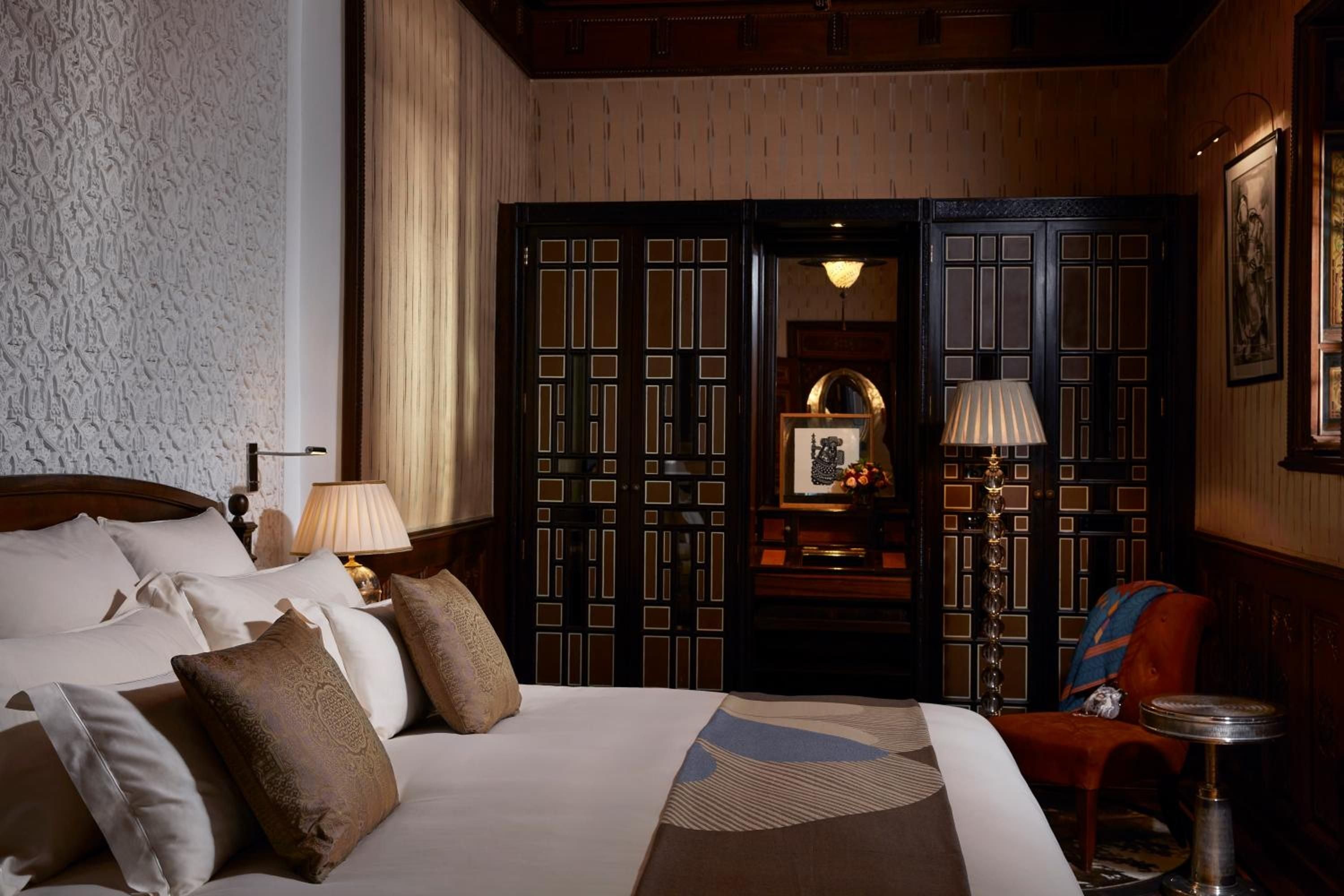 Intricate geometric woodwork frames a luminous alcove while silk lampshades cast golden warmth across crisp linens and hand-carved details in this palatial riad suite.