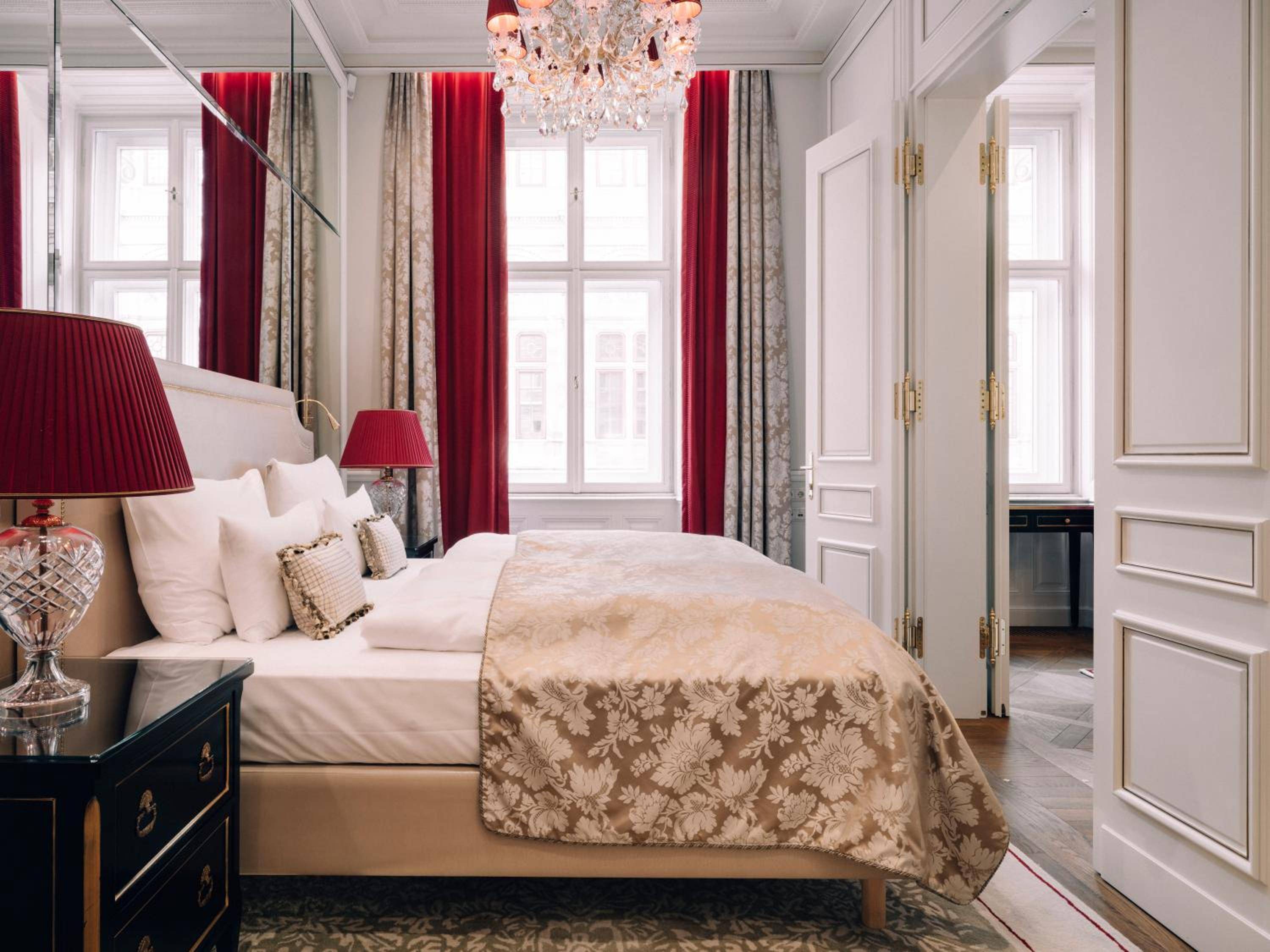 Crimson silk curtains frame tall windows while crystal chandeliers cast warm light across this opulent suite, where Viennese elegance meets sumptuous comfort in every gilded detail.