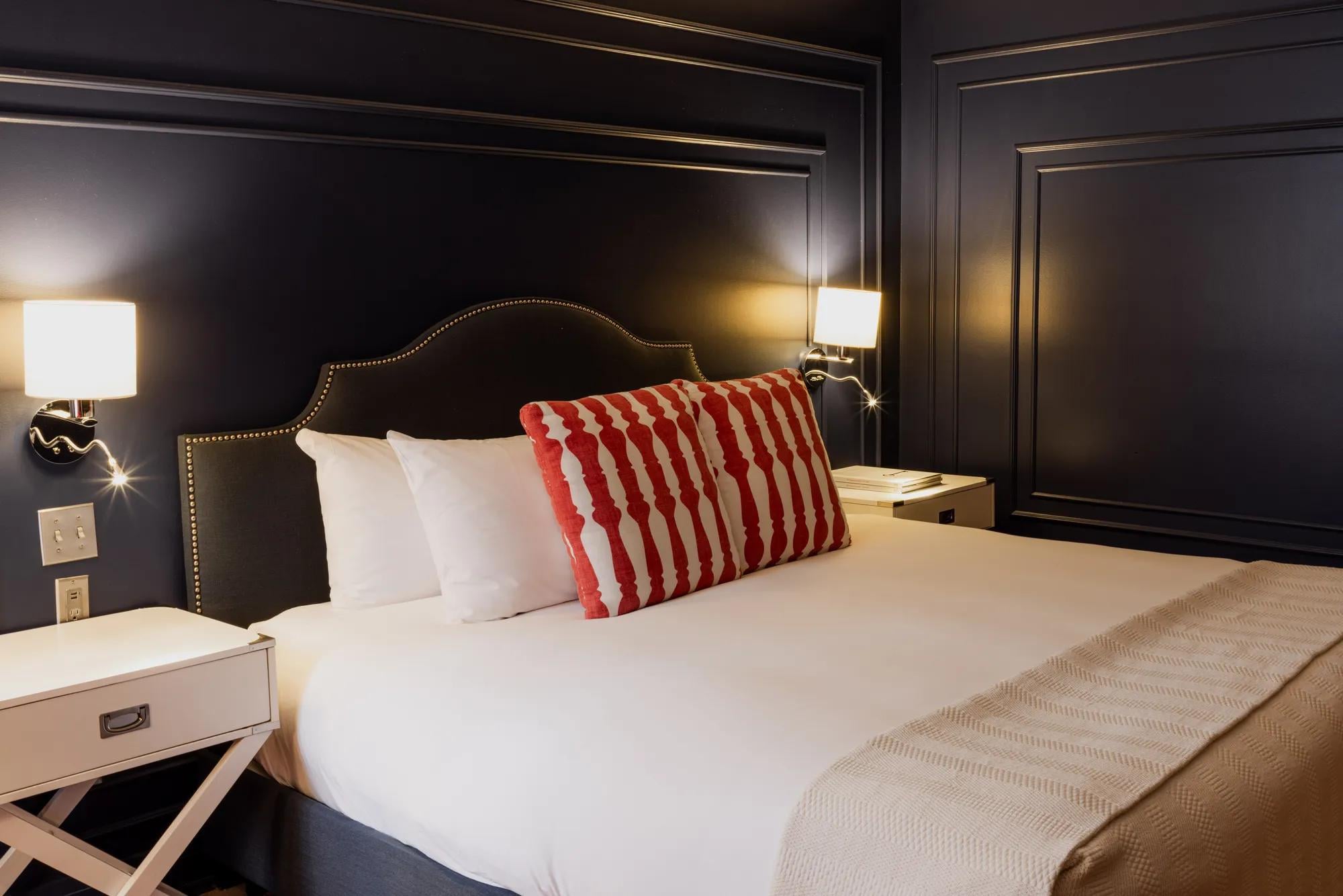 Dark paneled walls frame intimate sleeping quarters where brass nailhead trim catches lamplight, and a bold red geometric pillow punctuates crisp white linens.