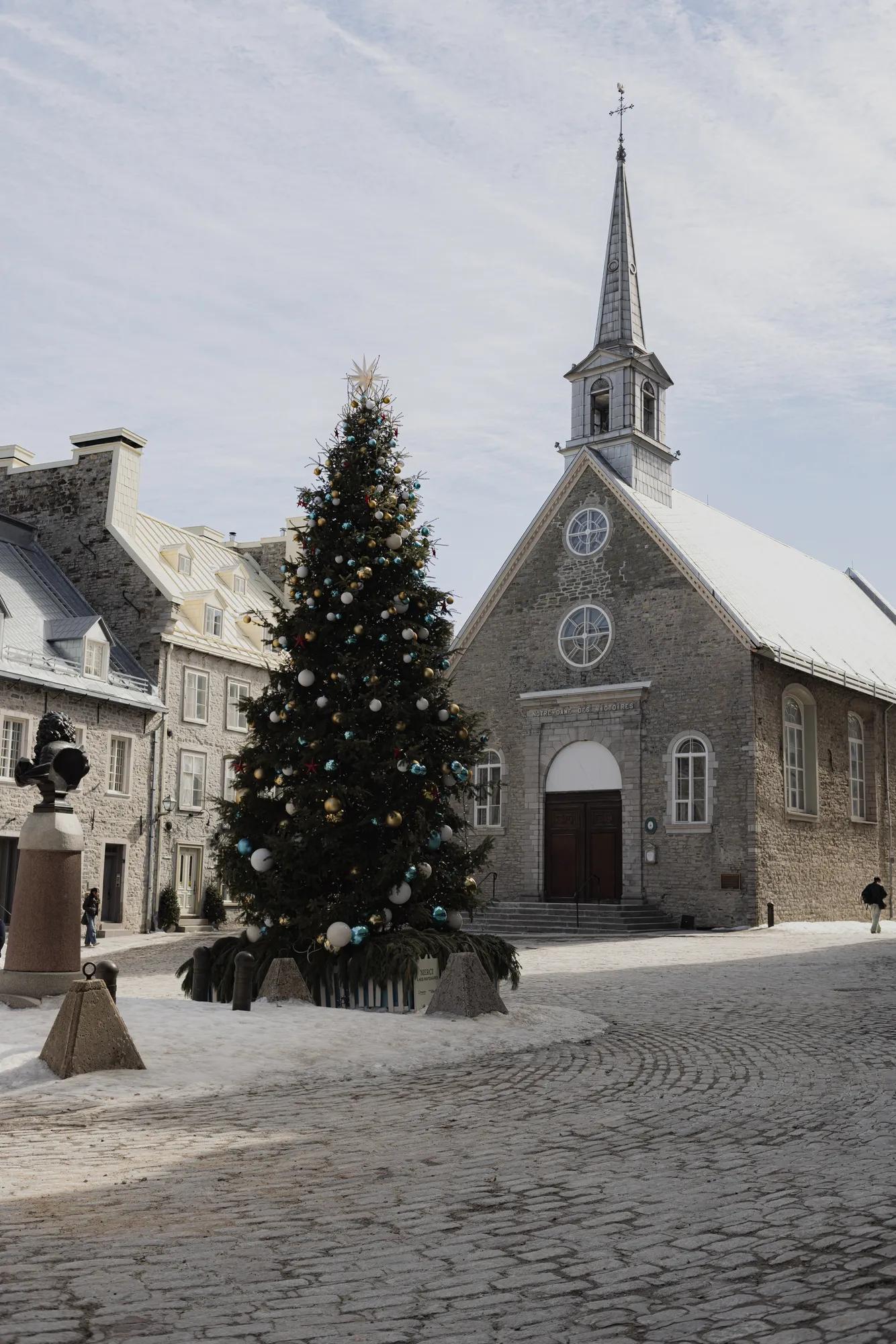 Snow dusts the cobblestones of Place Royale, where a festive Christmas tree stands before the historic Notre-Dame-des-Victoires church in Old Quebec's heart.