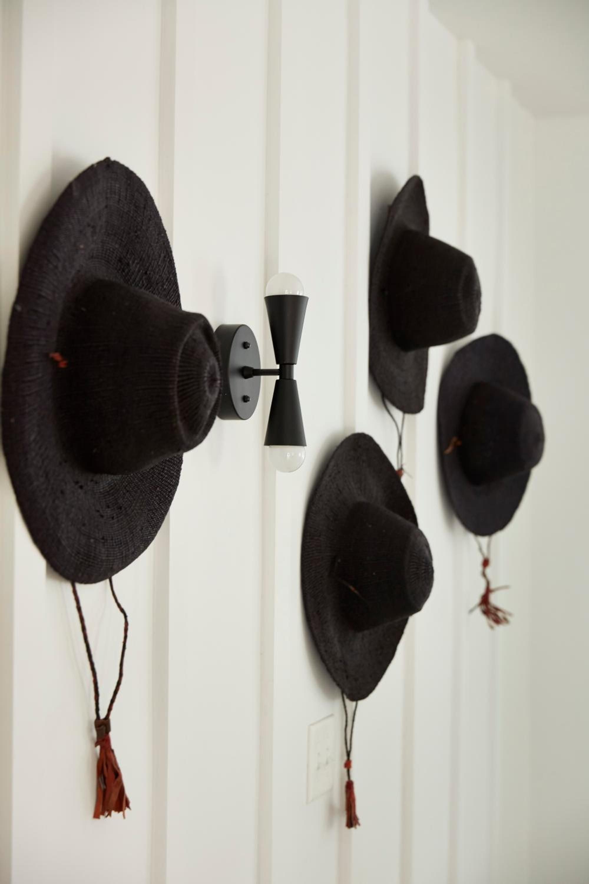 Black woven hats with crimson tassels create an artful display against white paneled walls, while a modern sconce adds geometric contrast to this thoughtful interior vignette.