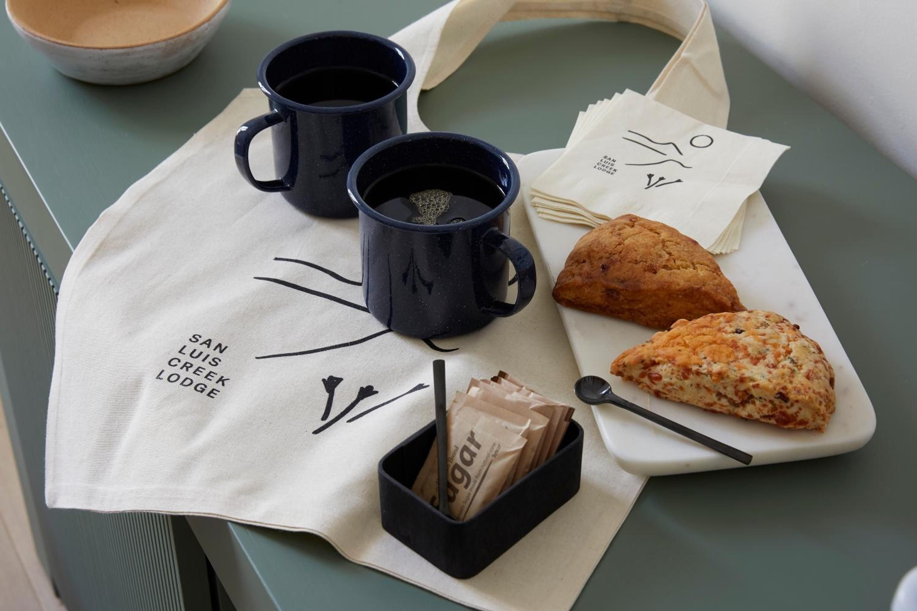Navy ceramic mugs cradle morning tea beside fresh-baked scones on rustic wooden boards, while branded canvas totes hint at the lodge's thoughtful hospitality touches.