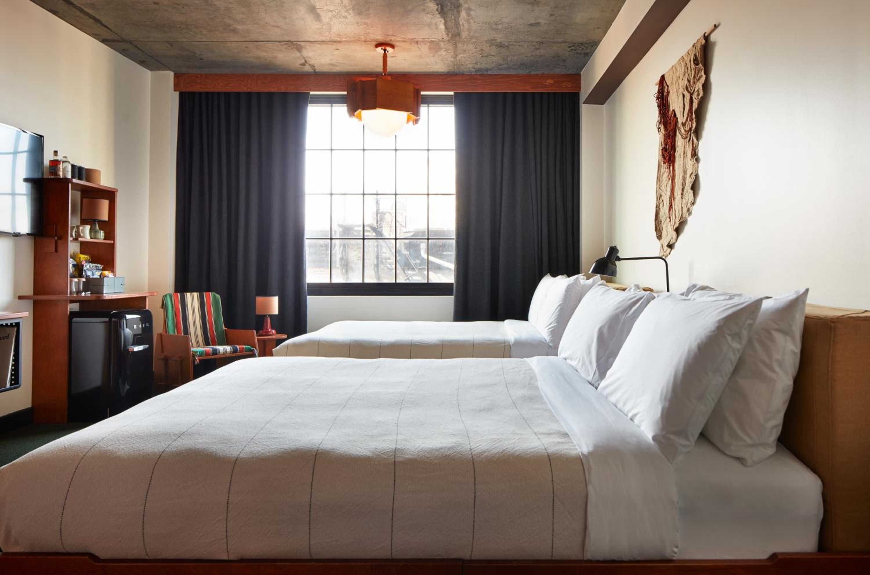 Raw concrete ceilings meet warm wood accents in thoughtfully designed guest rooms, where striped vintage chairs and macramé wall hangings create Brooklyn's signature creative energy.