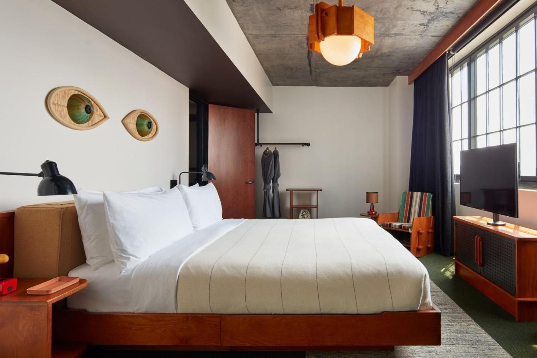 Warm wood tones and whimsical eye-shaped wall art create an inviting cocoon, where exposed concrete beams meet mid-century modern furnishings and crisp white linens.