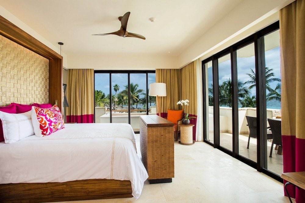 Vibrant magenta accents pop against crisp white linens while floor-to-ceiling windows frame swaying palms and turquoise Caribbean waters beyond the private terrace.