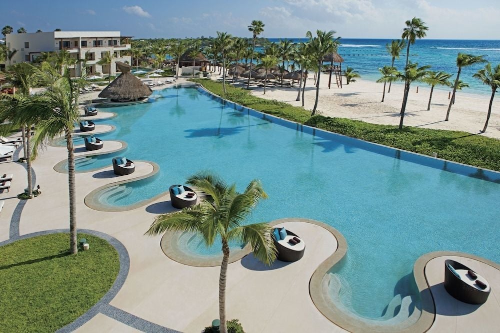 Infinity pools cascade toward white sand and turquoise Caribbean waters, while circular swim-up loungers create private floating sanctuaries beneath swaying palms.