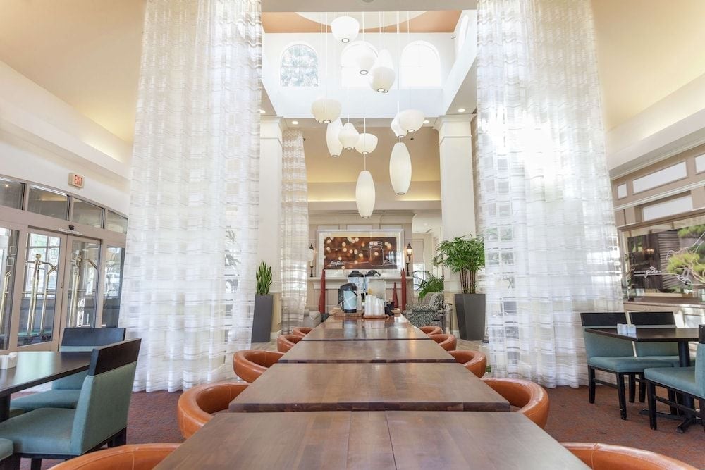 Floating white curtains frame a communal dining space where warm orange chairs invite intimate conversations beneath sculptural pendant lights and soaring ceilings.