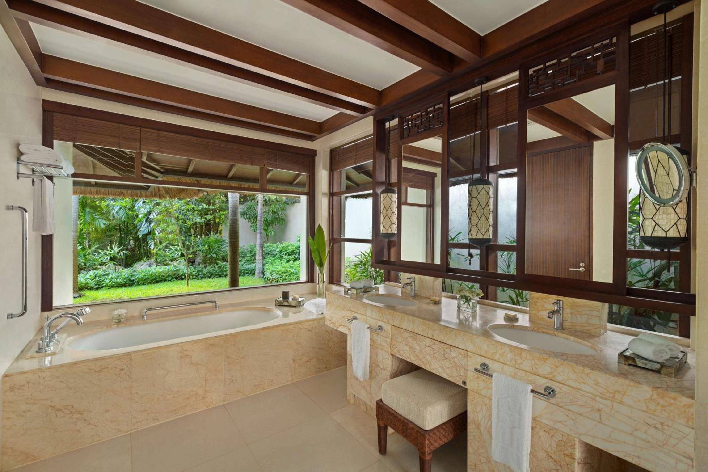 Warm teak beams frame marble vanities and a soaking tub with garden views, where tropical breezes drift through latticed windows in this sanctuary-like bathroom.