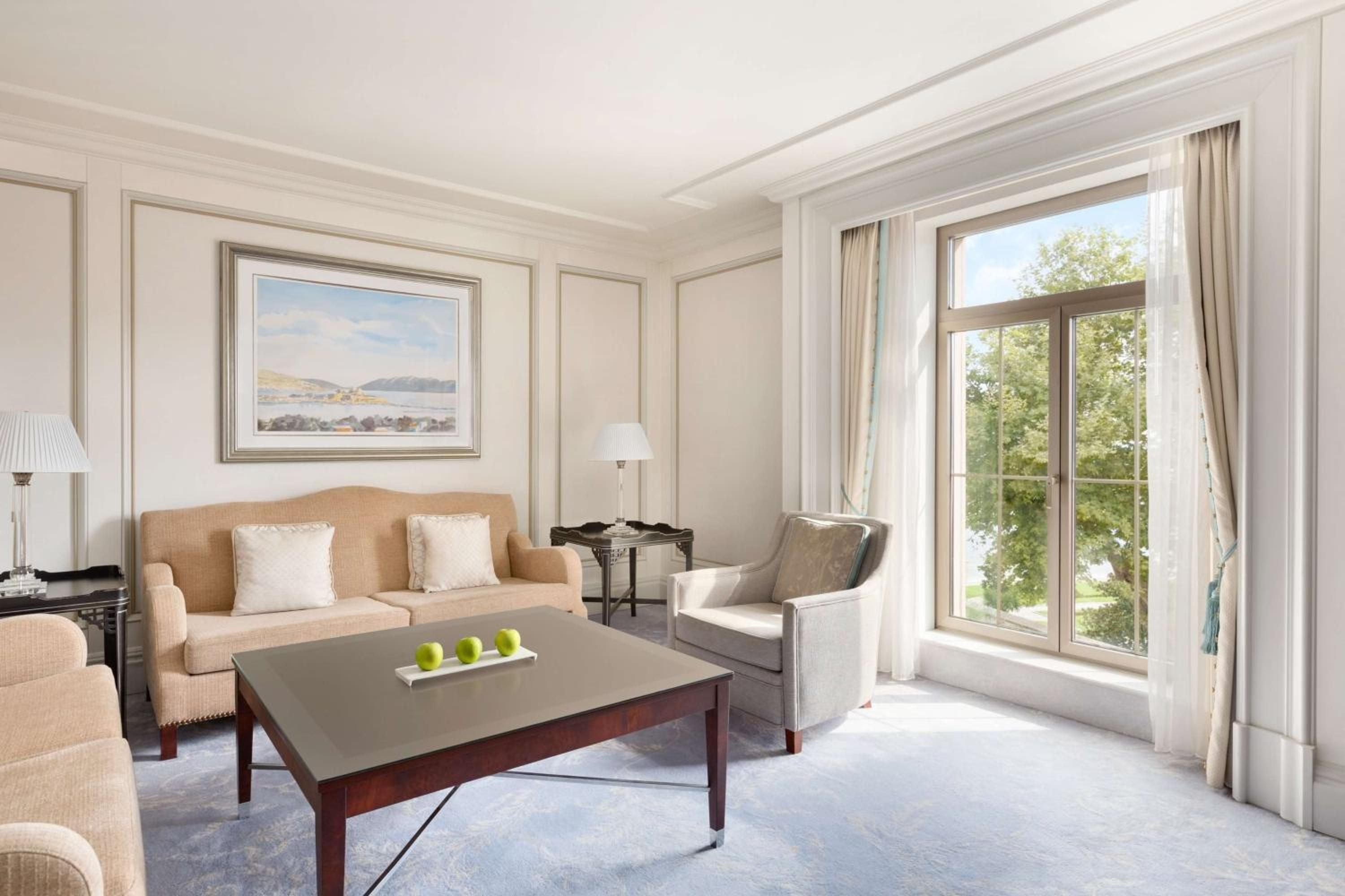 Soft natural light filters through floor-to-ceiling windows in this elegantly appointed suite, where neutral tones and classic furnishings create a serene Istanbul sanctuary overlooking verdant gardens.