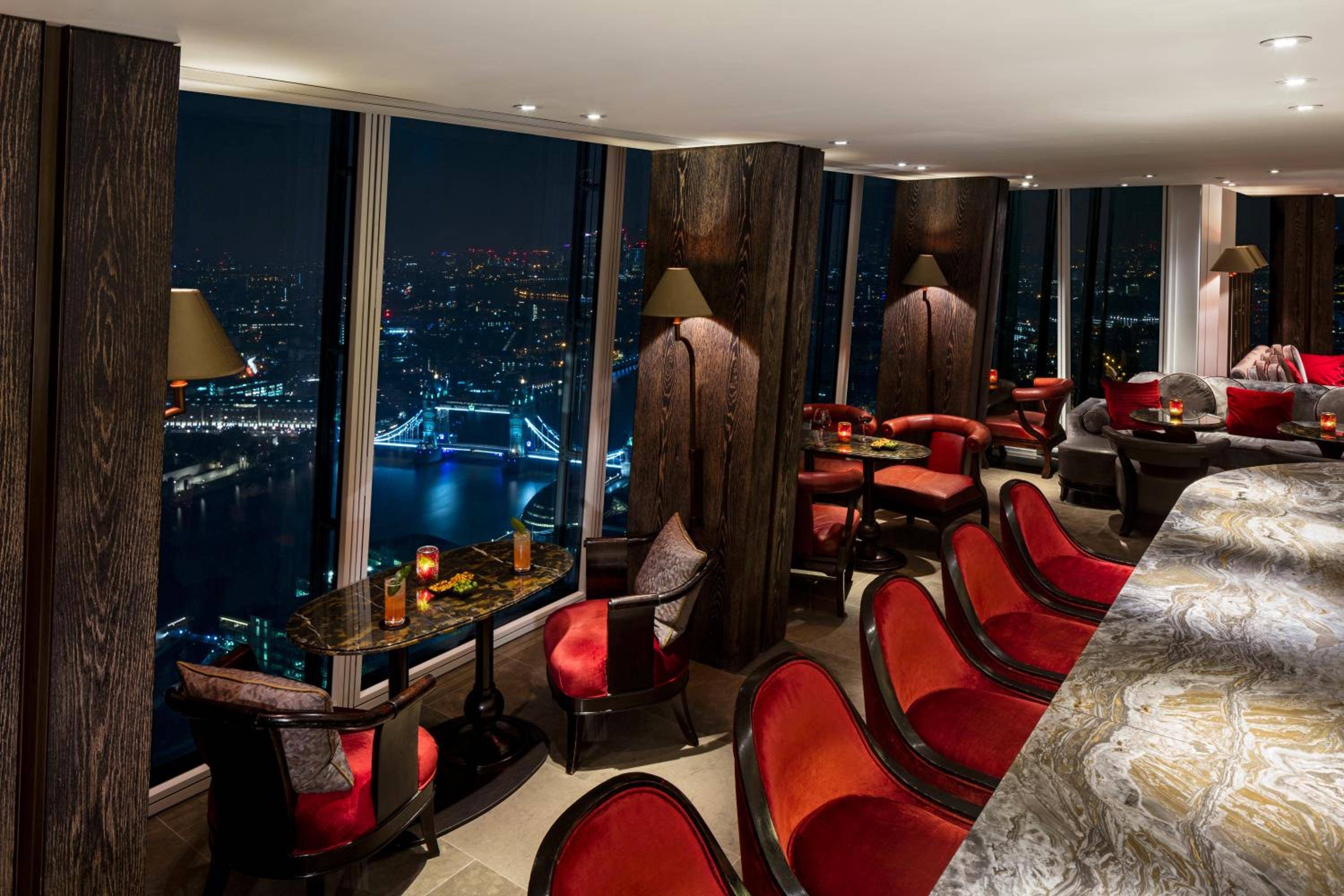 Crimson velvet chairs frame Tower Bridge's illuminated arches through floor-to-ceiling windows, while amber candlelight creates an intimate atmosphere high above London's glittering sprawl.