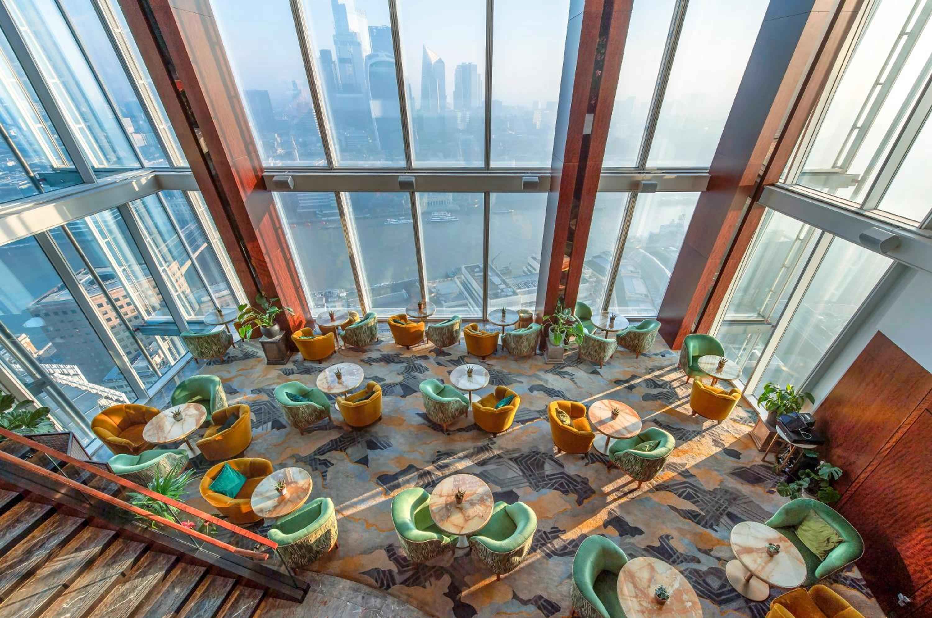 Jewel-toned velvet chairs create intimate conversation circles beneath soaring glass walls, where London's skyline stretches endlessly beyond this elevated sanctuary.