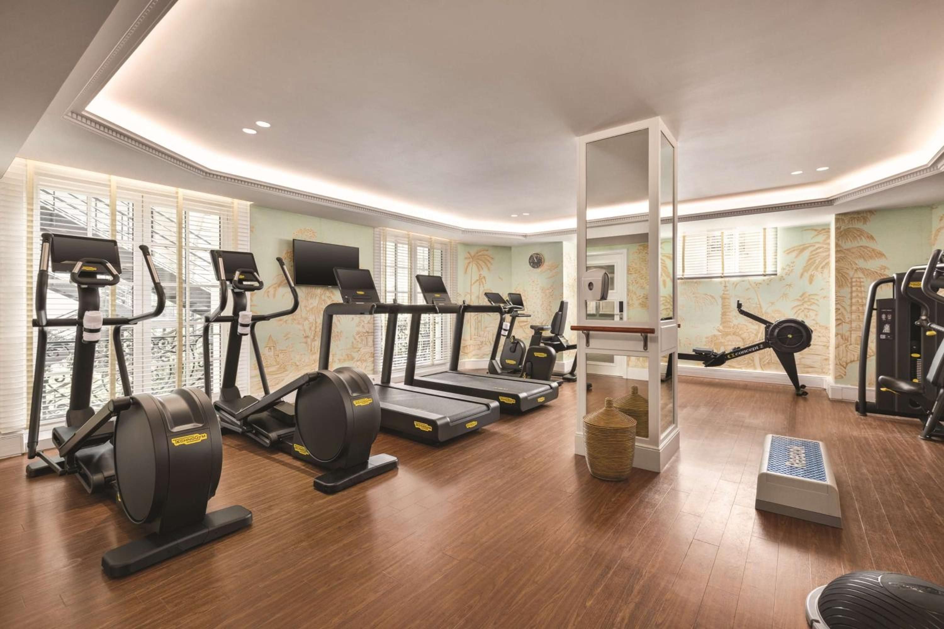 State-of-the-art cardio equipment lines warm hardwood floors beneath elegant coffered ceilings, where tropical murals and natural light create an unexpectedly serene workout sanctuary.