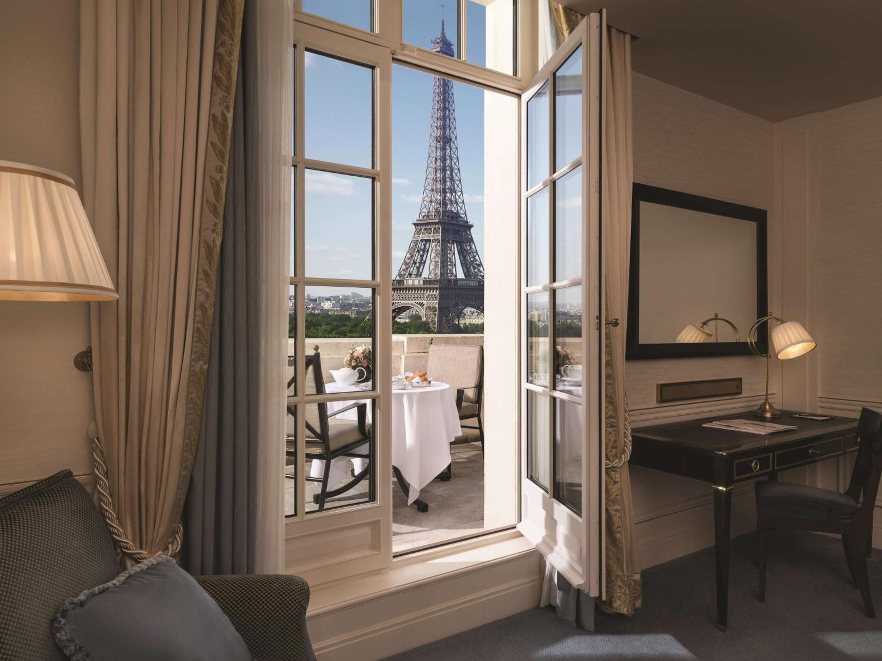 French doors frame the iconic Eiffel Tower from this elegantly appointed guest room, where afternoon light streams across polished wood and classic Parisian details.
