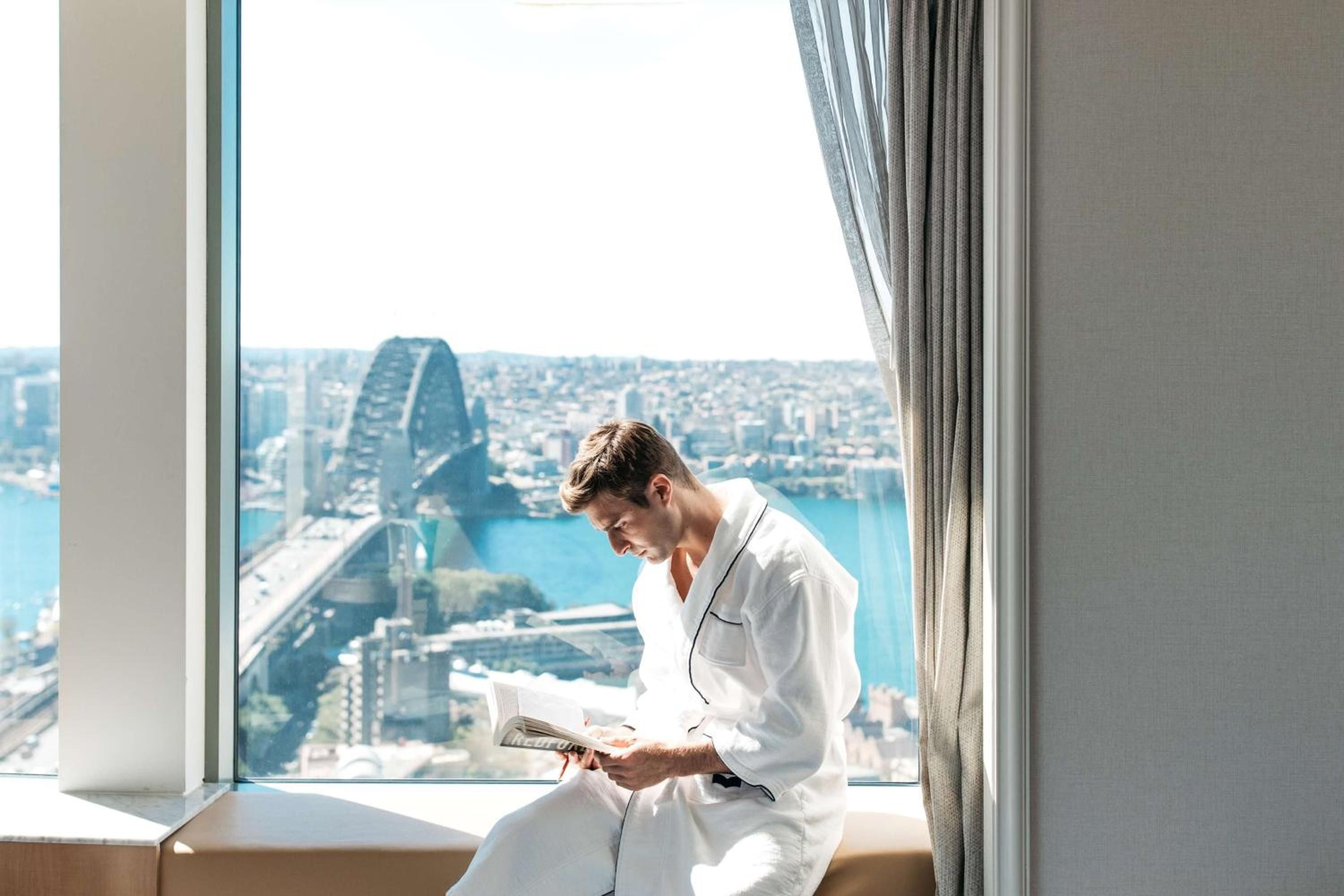 Morning light streams through floor-to-ceiling windows as guests savor coffee and newspapers, with Sydney Harbour Bridge creating a stunning backdrop from this harbourside sanctuary.