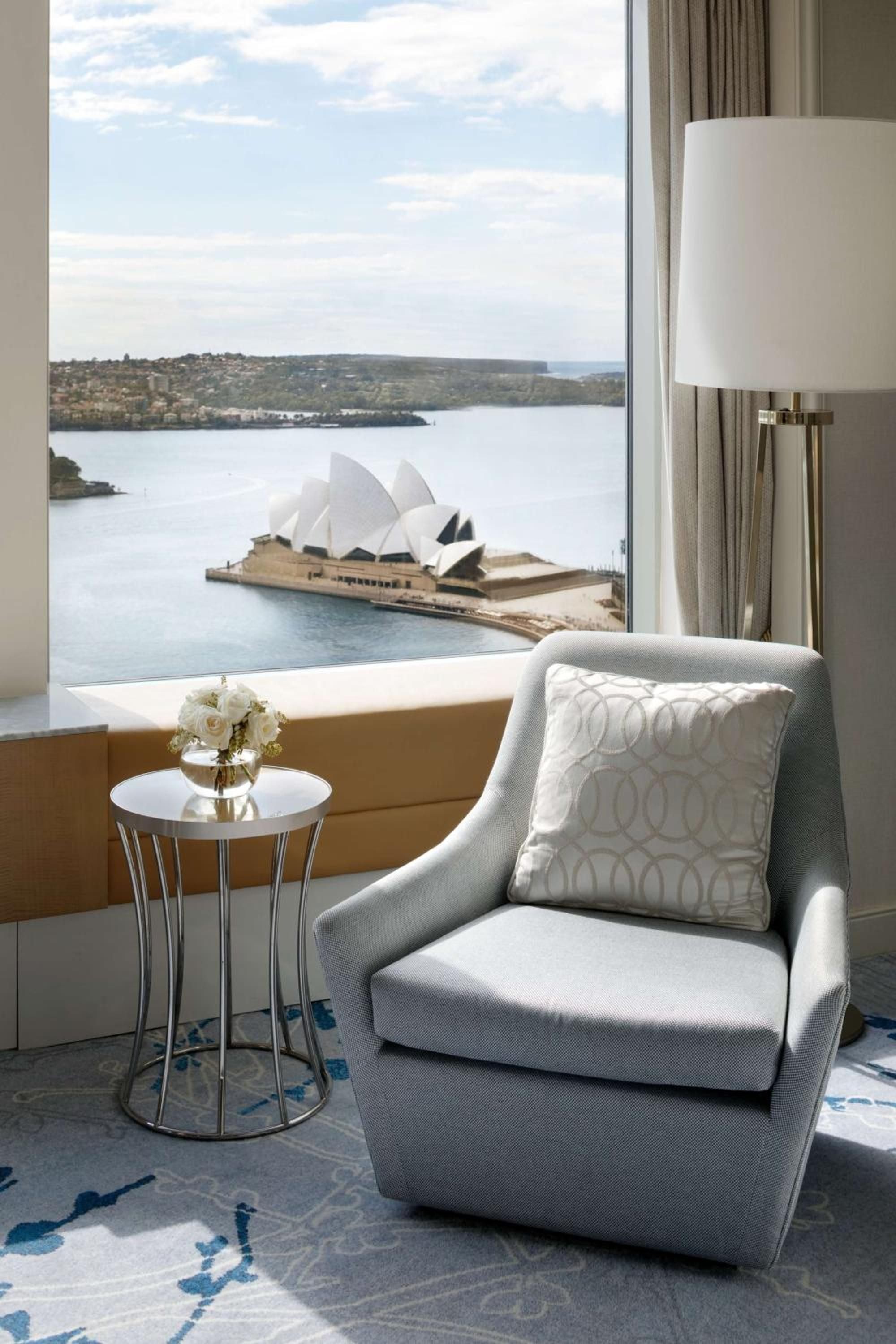 Plush armchair and metallic side table frame stunning harbor views, where Sydney Opera House's iconic shells gleam across sparkling blue waters from this coveted corner suite.