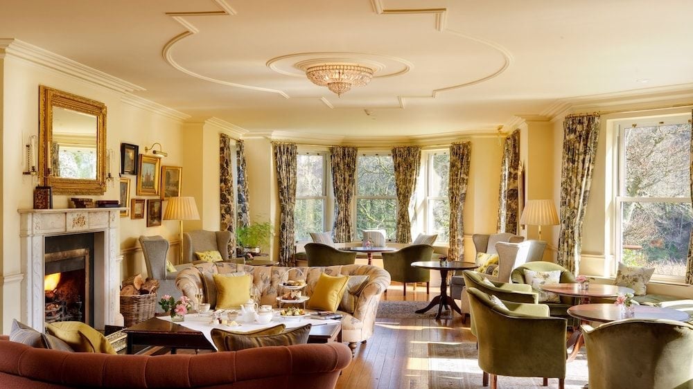Sumptuous velvet armchairs and flickering firelight create an intimate drawing room where afternoon tea unfolds beneath an ornate crystal chandelier and coffered ceiling.