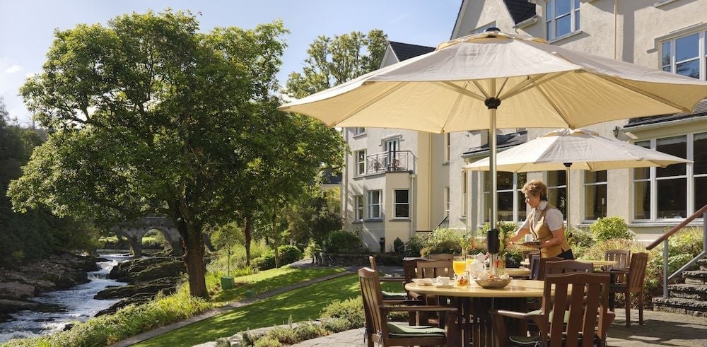 Riverside dining unfolds beneath cream umbrellas on the terrace, where the gentle murmur of flowing water mingles with clinking glasses and dappled sunlight through ancient trees.