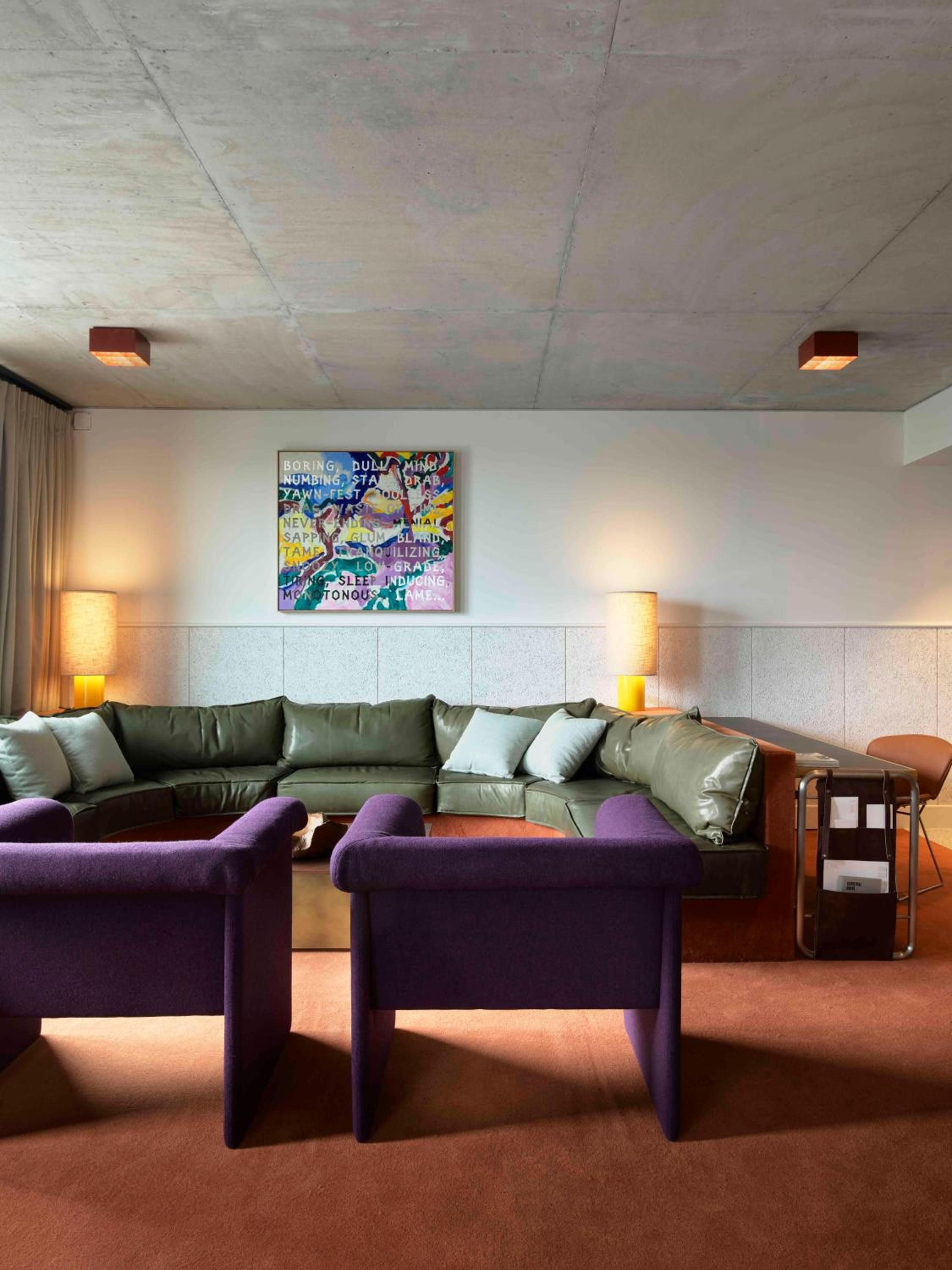 Olive leather sectionals and plush purple ottomans create intimate conversation nooks beneath exposed concrete ceilings, where warm amber lighting softens industrial edges with residential comfort.