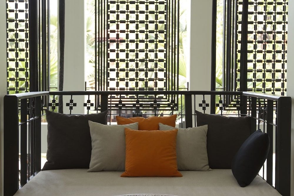 Geometric shadows dance through intricate latticed screens onto plush daybeds adorned with burnt orange pillows, creating an intimate reading nook bathed in filtered tropical light.