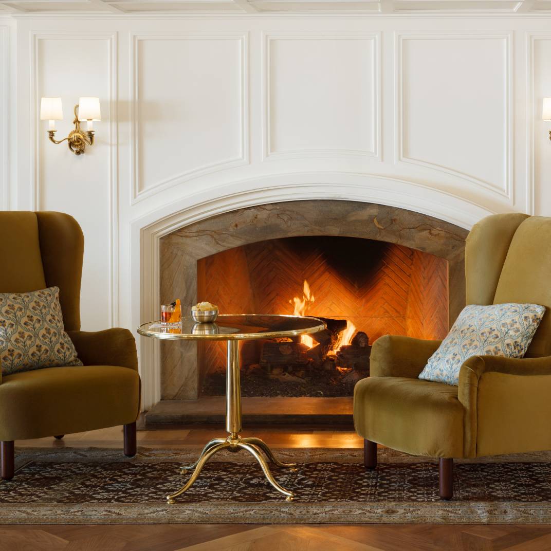 Golden flames dance behind herringbone brick as wingback chairs invite intimate conversation over evening cocktails in this perfectly appointed corner sanctuary.