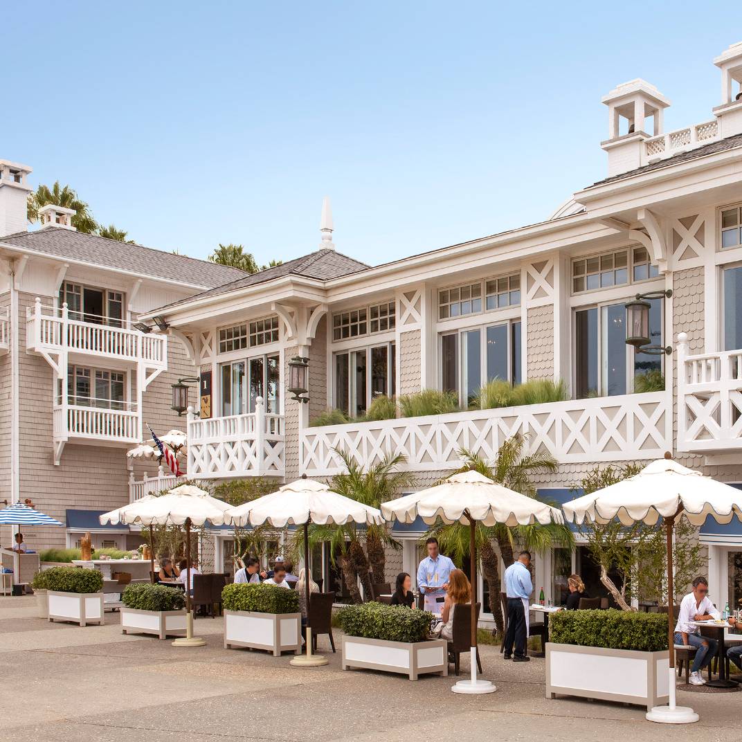 Cream umbrellas shade intimate terrace tables where servers weave between guests savoring coastal cuisine, while weathered shingle cottages frame this quintessential Santa Monica dining scene.