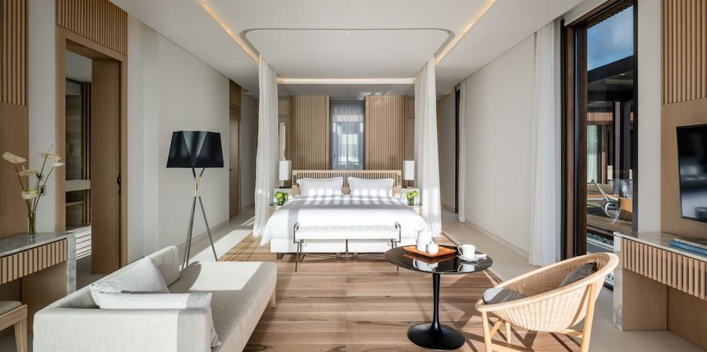 Four-poster beds draped in crisp white linens anchor these airy suites, where warm wood accents and floor-to-ceiling windows create an understated sanctuary.