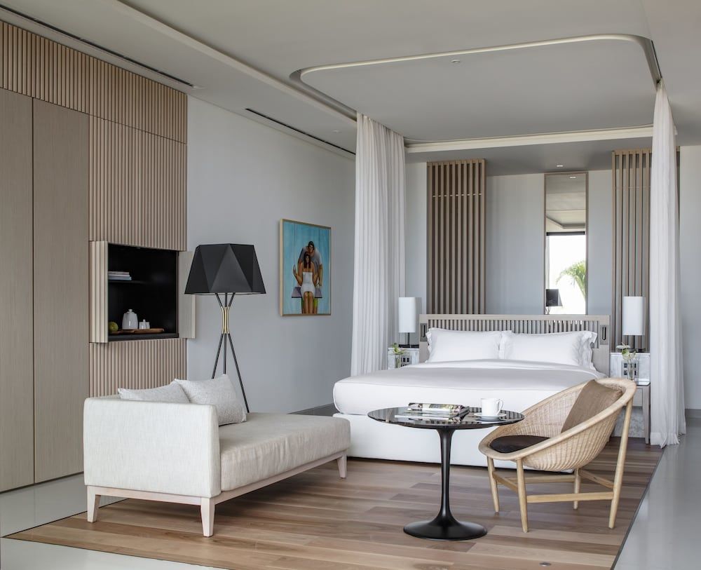 Crisp white linens frame the four-poster bed while warm wood slats create intimate alcoves in this sophisticated suite where modern luxury meets tropical serenity.