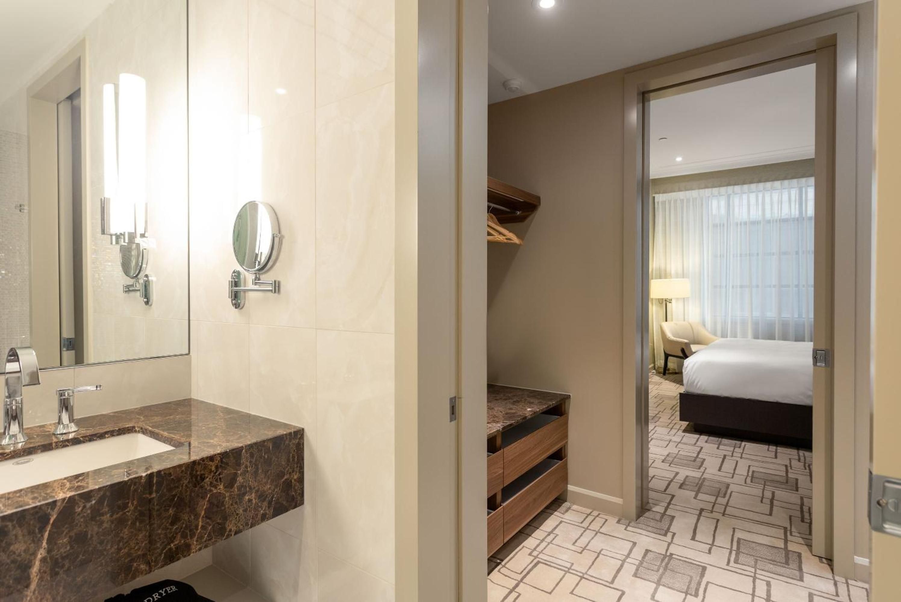 Marble vanity meets warm wood closet storage in this thoughtfully designed suite, where natural light streams through geometric carpeting toward the inviting bedroom beyond.