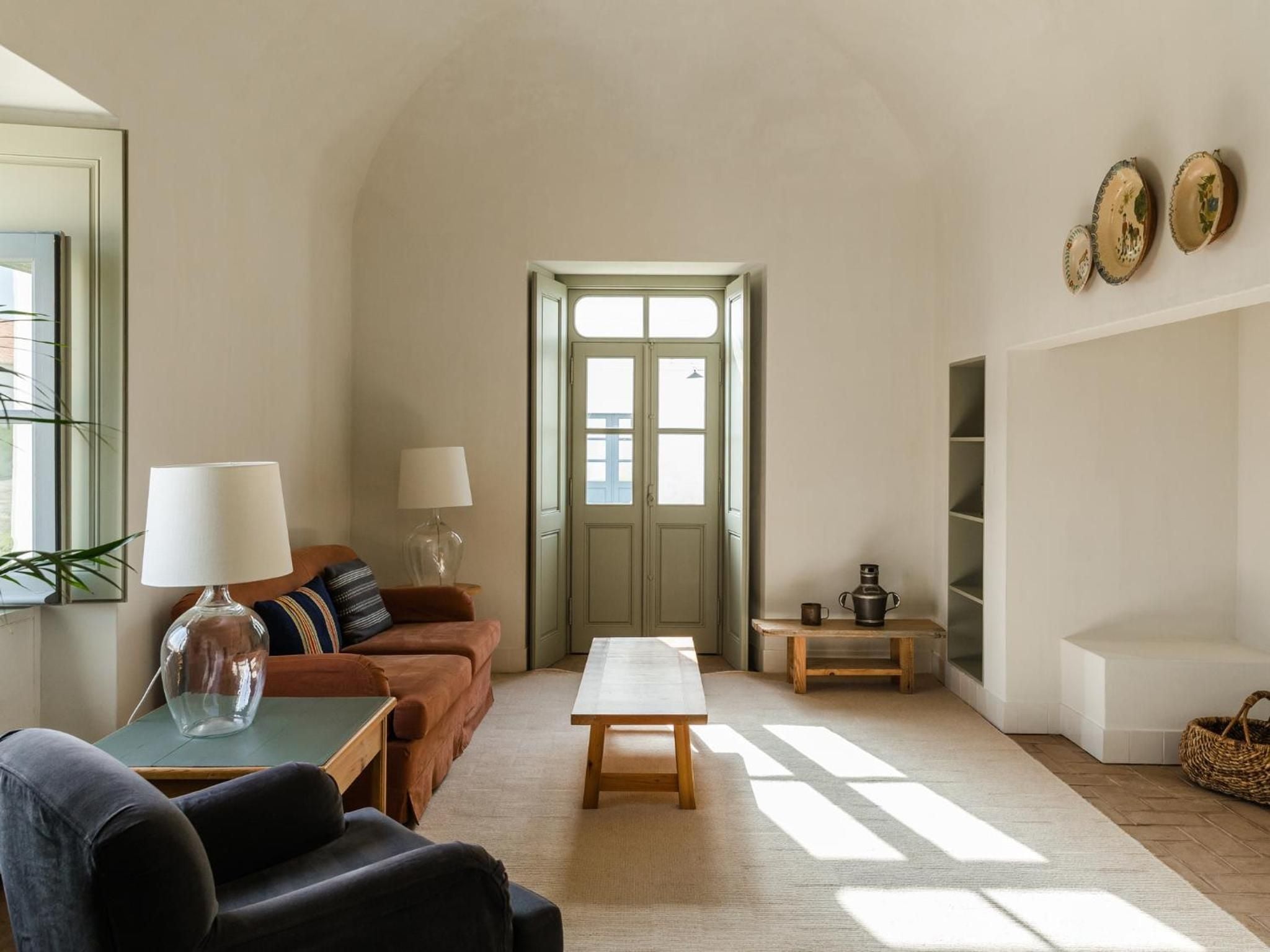 Sunlight streams through sage-green French doors into this serene living space, where burnt orange velvet meets neutral tones and handcrafted ceramics create an effortlessly sophisticated retreat.