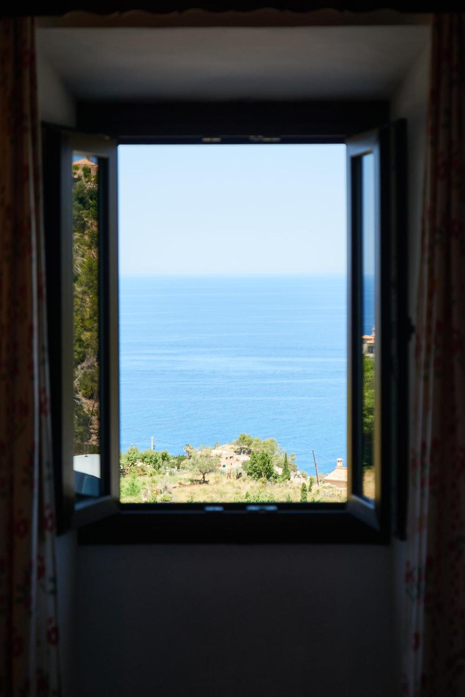 Open shutters frame a postcard-perfect vista of azure Mediterranean waters and terraced hillsides dotted with ancient stone dwellings from this intimate guest room.