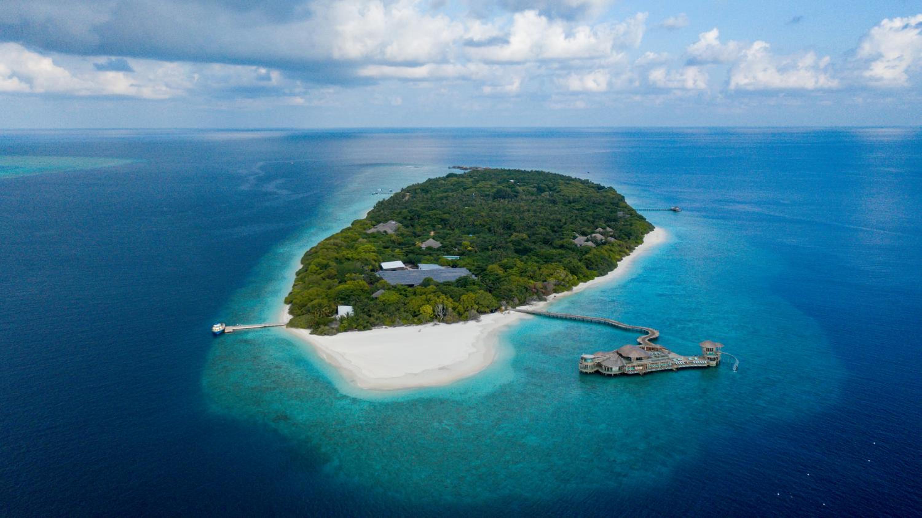 Pristine white sand embraces this jewel-like atoll, where overwater villas and dense tropical canopy create an untouched paradise suspended in turquoise infinity.