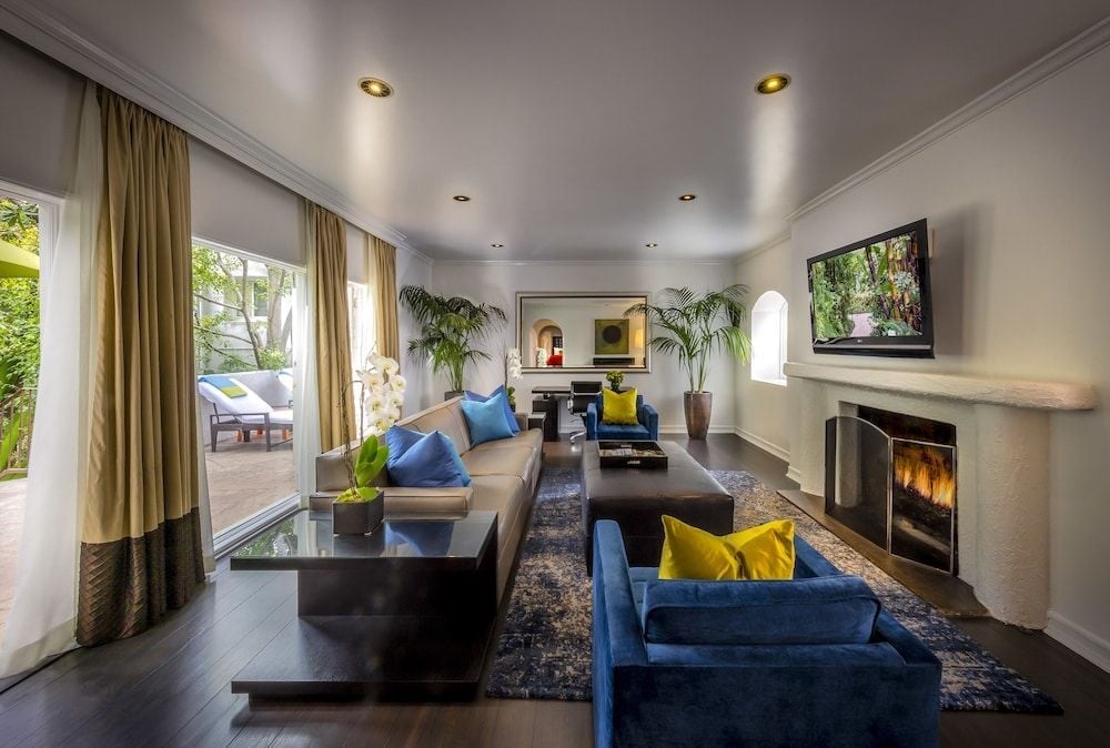 Recessed lighting casts a warm glow over plush blue velvet seating and dark wood floors, while French doors reveal a private terrace beyond flowing curtains.
