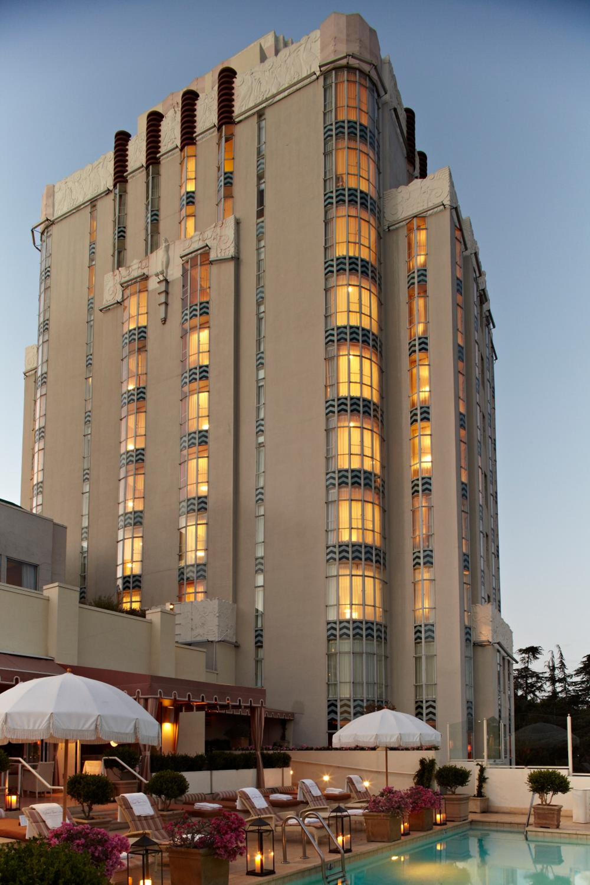 Golden hour illuminates the Art Deco tower's distinctive vertical windows while poolside umbrellas and flickering lanterns create an intimate terrace sanctuary beneath the Hollywood Hills.