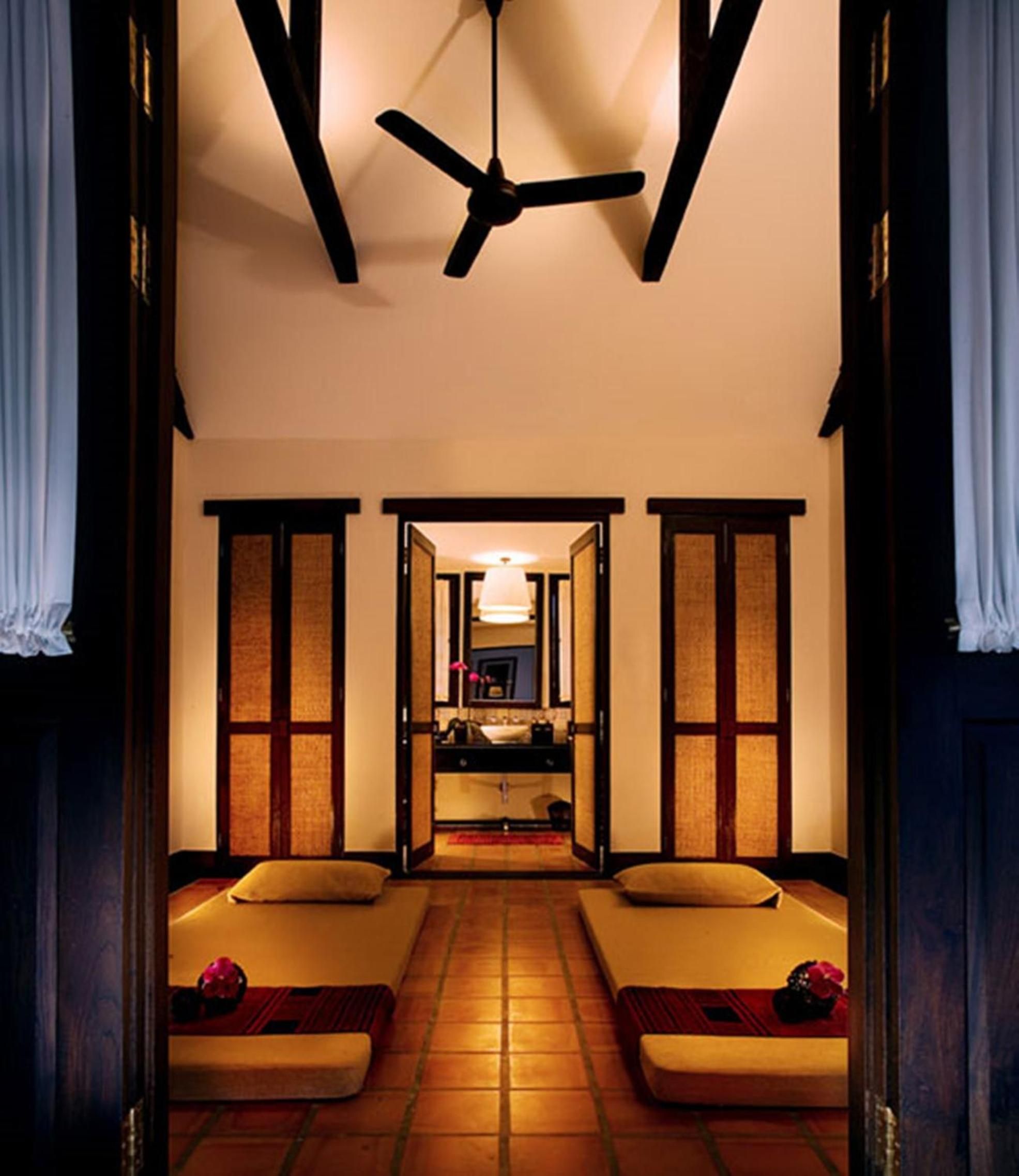 Twin massage beds await beneath exposed wooden beams and traditional screens, where warm terracotta tiles and flickering ceiling fans create an intimate sanctuary for couples treatments.