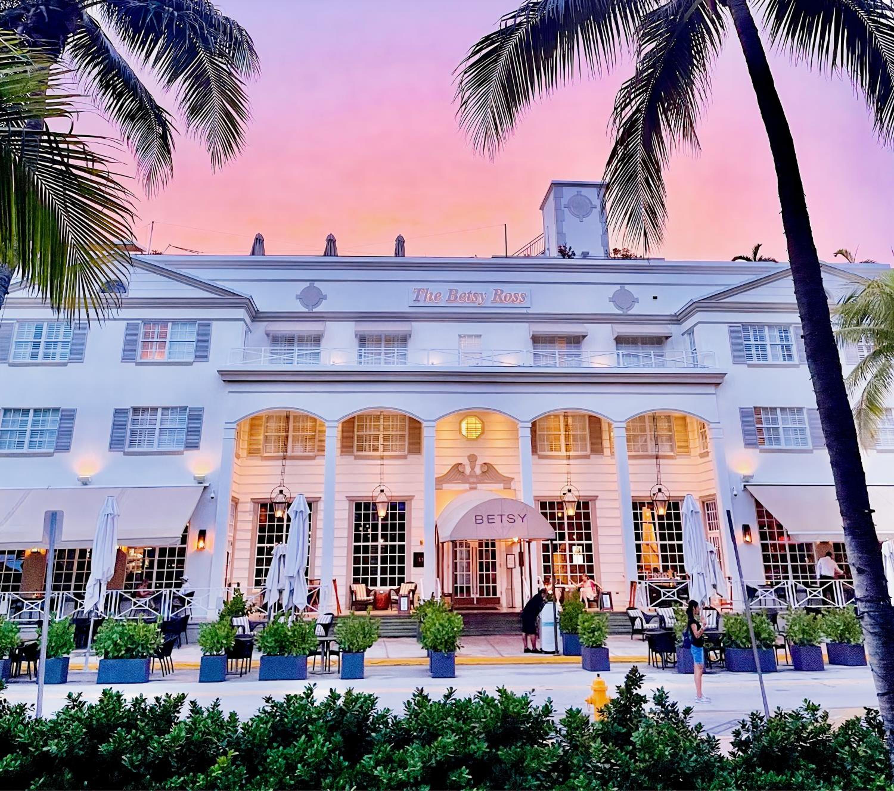 Palm fronds frame the Betsy Ross's elegant white facade as golden hour light bathes its colonial-inspired arches and outdoor dining terraces in warm coral hues.