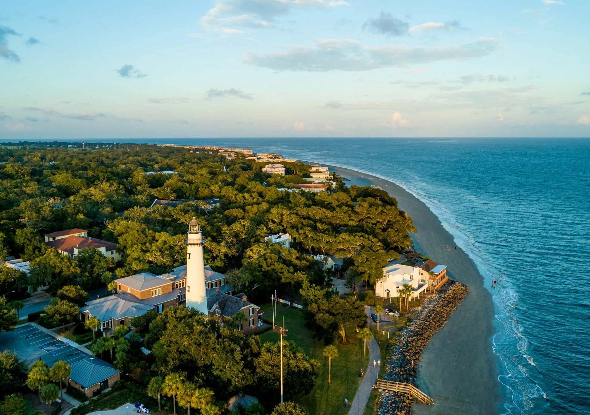 Golden hour light bathes the historic lighthouse and coastline, where palm-dotted grounds meet pristine sand and rolling Atlantic waves create an endless azure horizon.