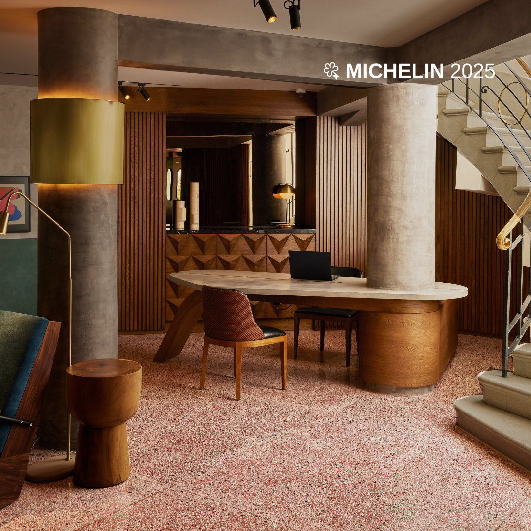Warm terrazzo floors and sculptural concrete columns frame the sophisticated lobby, where mid-century furniture mingles with geometric wood paneling beneath dramatic track lighting.