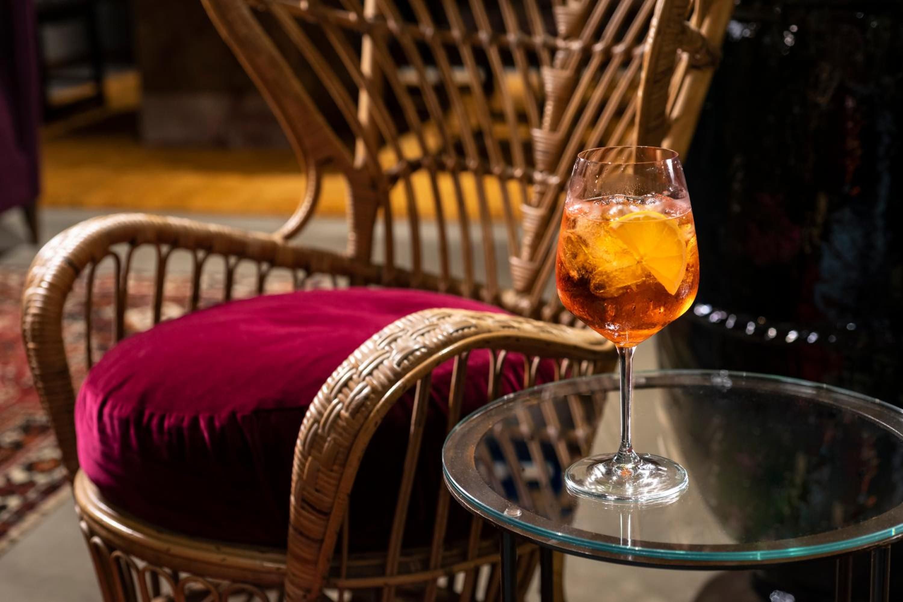 Amber aperitivo catches golden hour light on a glass table, surrounded by woven rattan chairs with jewel-toned cushions in this intimate corner.