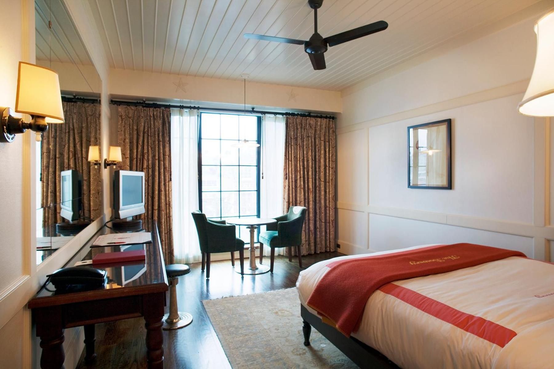 Exposed white beams frame intimate seating by tall windows, while rich burgundy accents warm the sophisticated guest room's clean-lined aesthetic.
