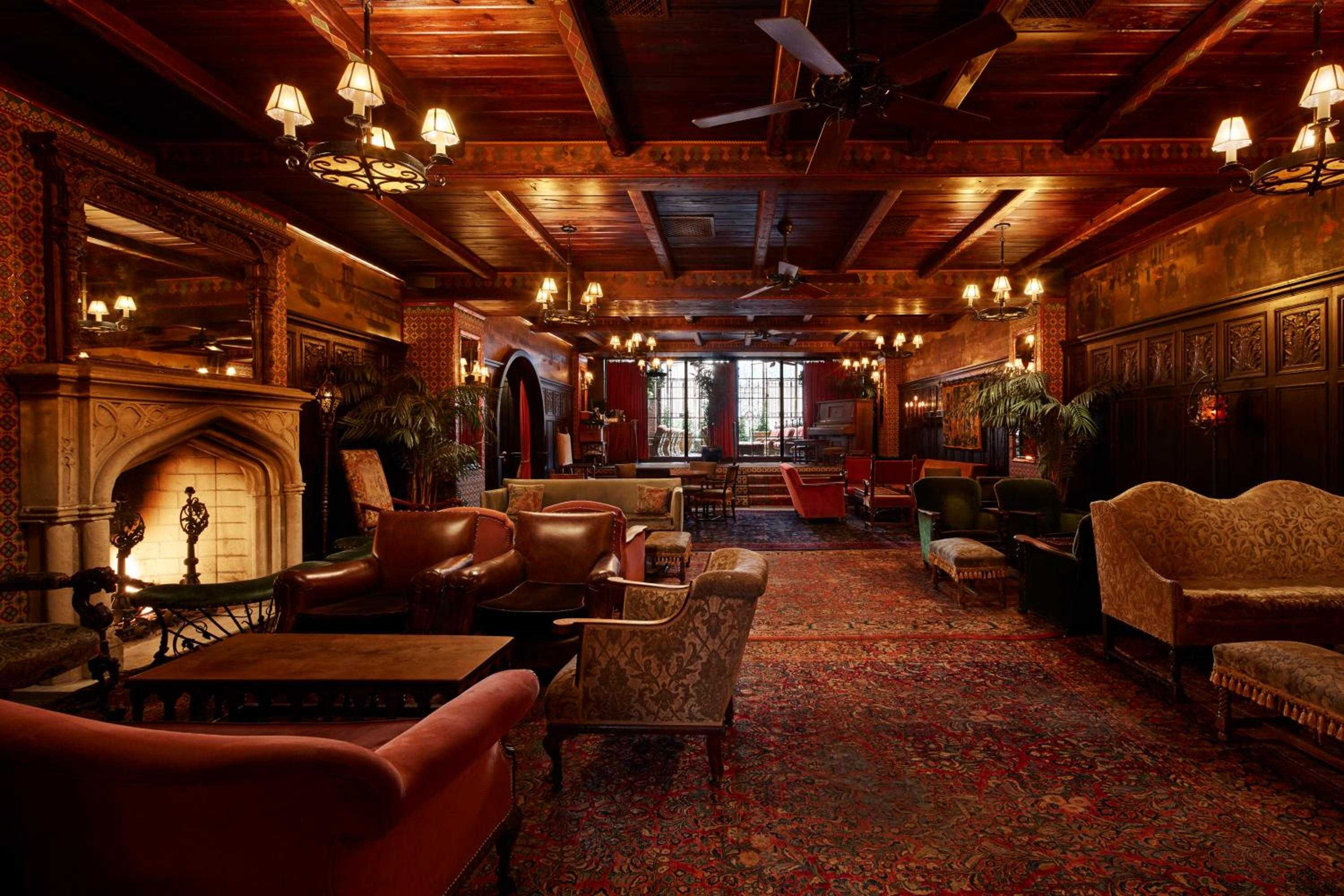 Exposed timber beams and wrought-iron chandeliers frame plush leather seating around a carved stone fireplace, creating an intimate English manor atmosphere.