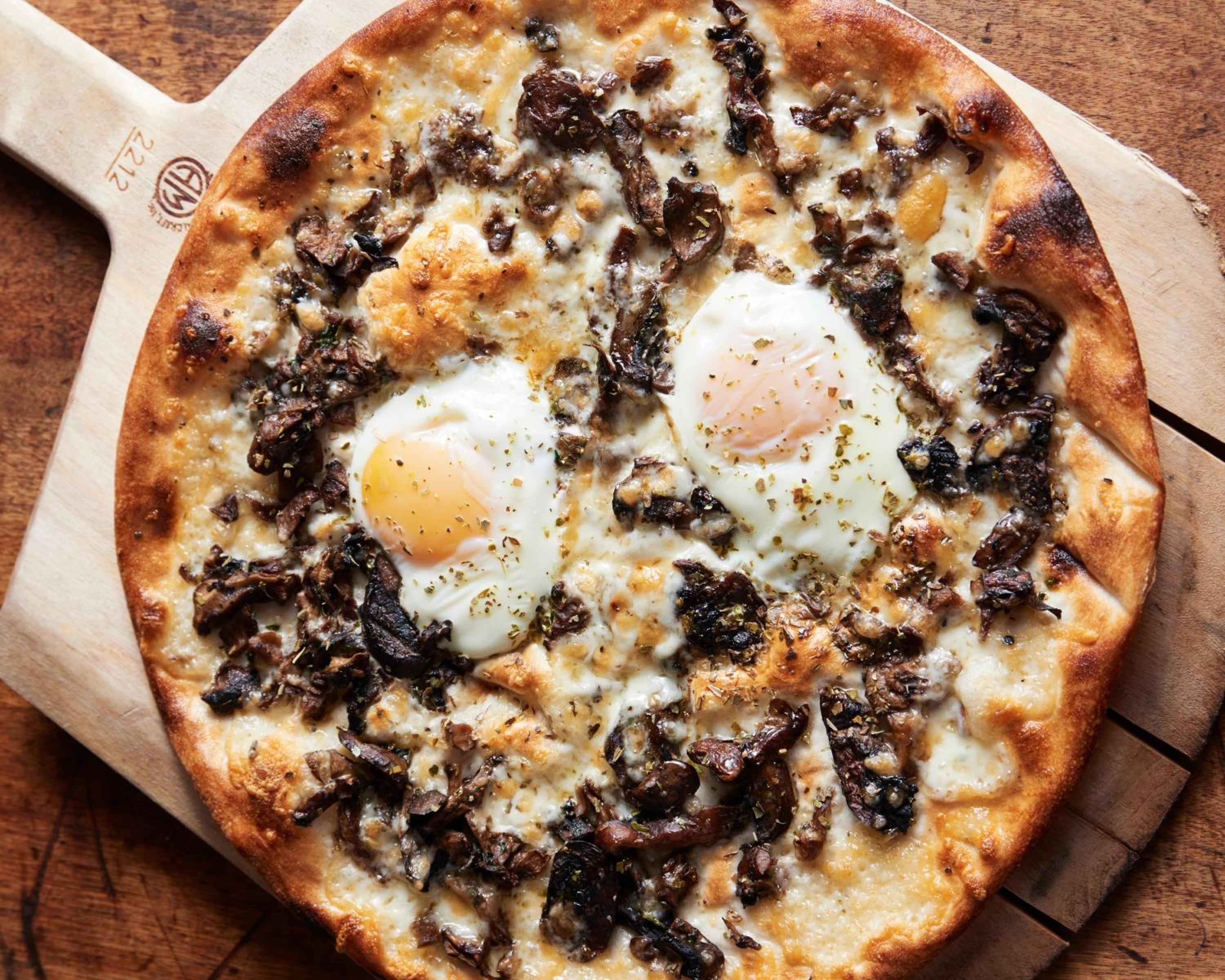 Farm-fresh eggs crown a rustic pizza topped with wild mushrooms and caramelized onions, served on charred wood that still whispers of the oven's heat.