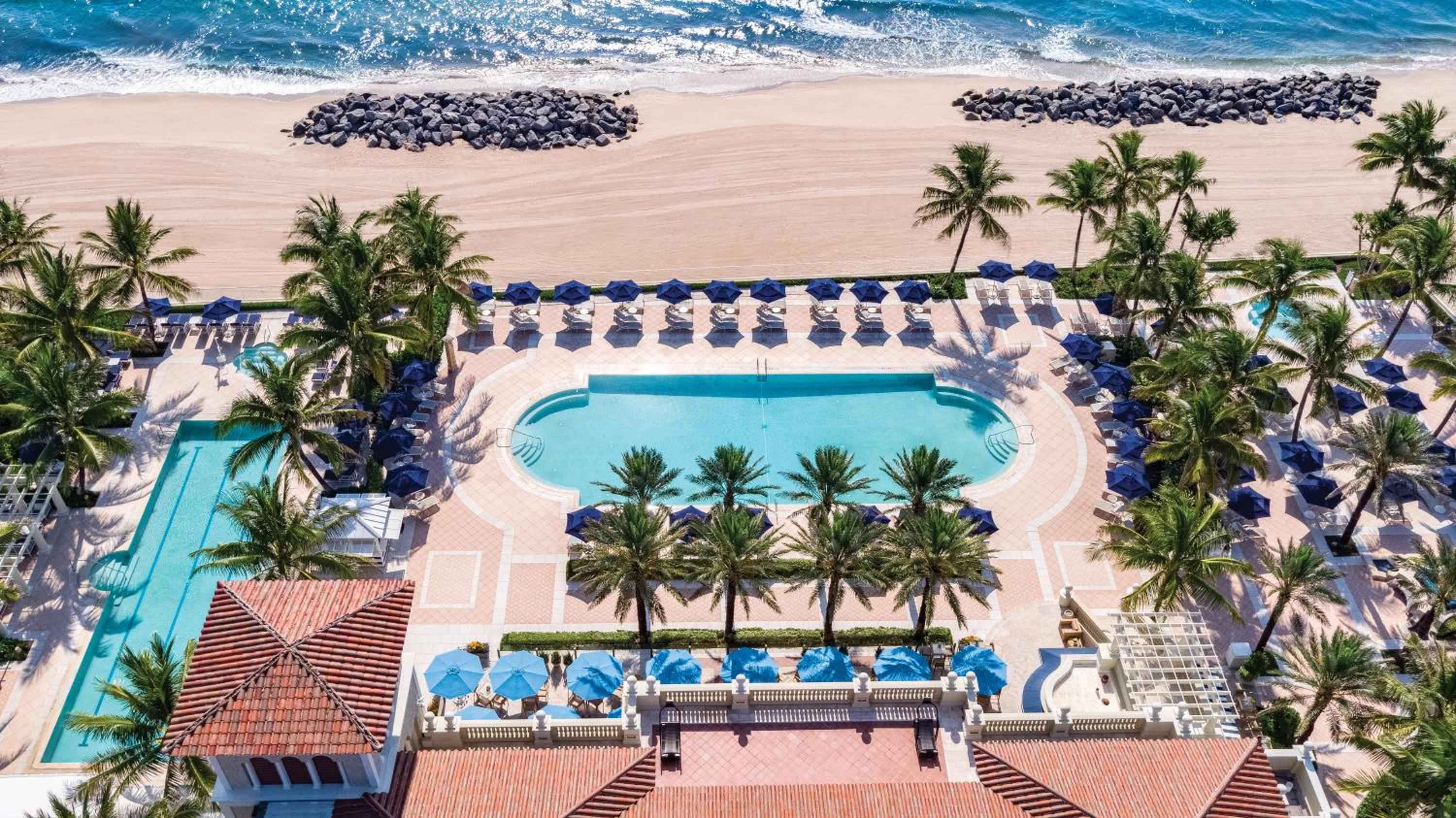 Twin pools cascade toward pristine sand where azure waves meet carefully arranged stone breakwaters, creating an oceanfront sanctuary framed by swaying palms and blue umbrellas.